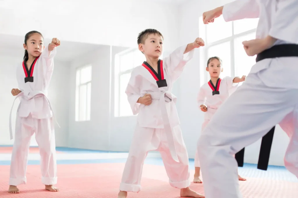 Crianças praticando karatê em aula de artes marciais, com instructor ensinando técnica a alunos adolescentes em ginásio bem iluminado.
