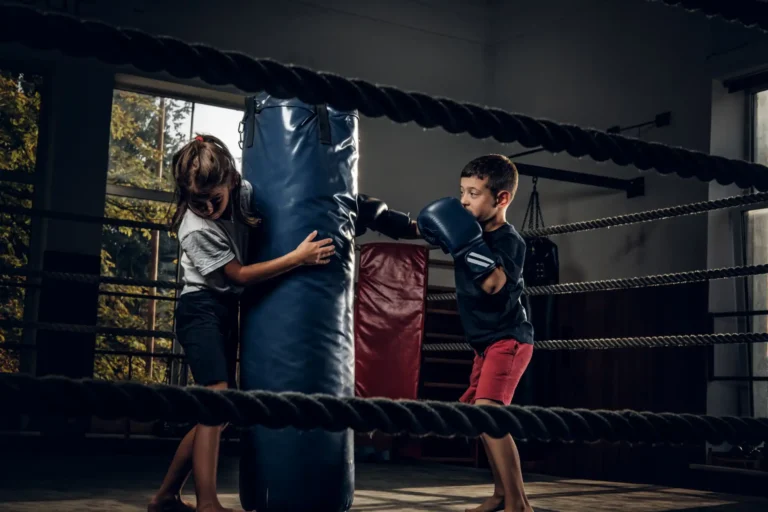 Duas crianças treinando boxe na academia, com um menino lançando golpes e uma menina apoiando-se na almofada de treino, ambiente de luta com iluminação natural.