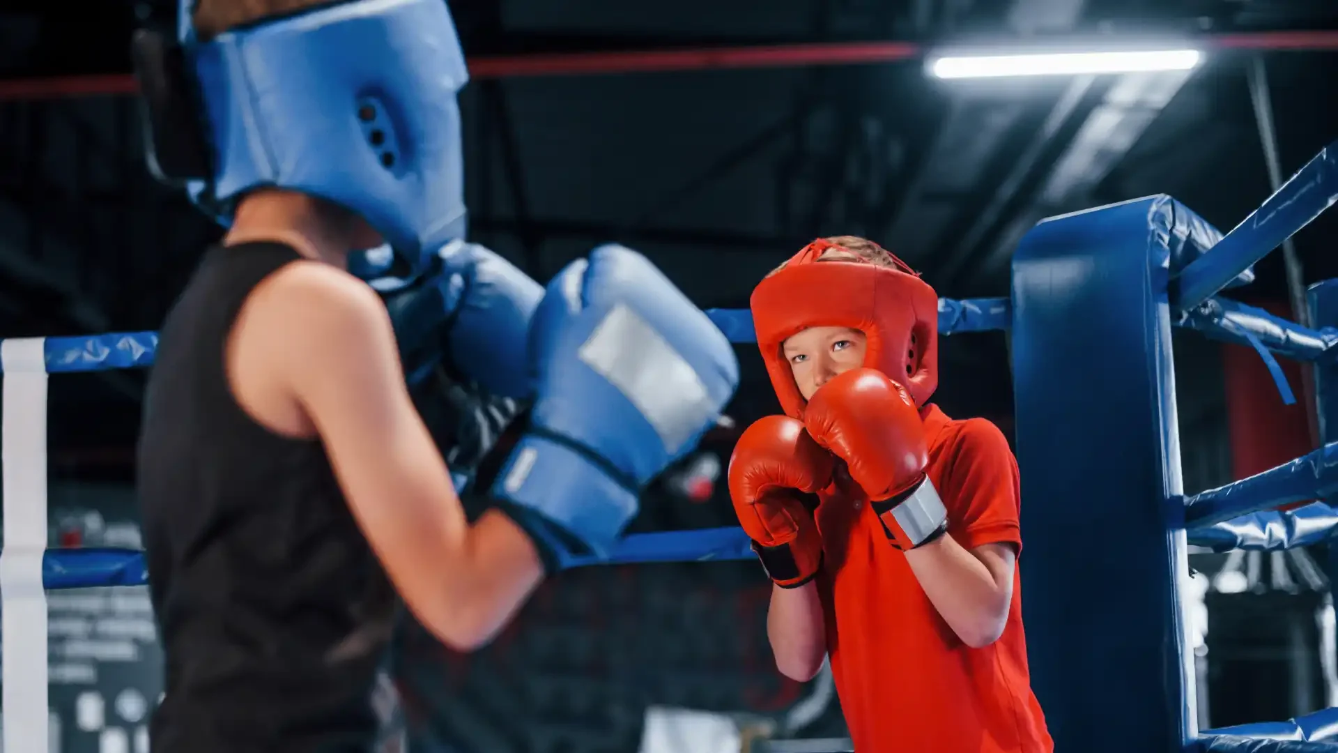 Duelo de crianças em treino de boxe, com um menor em equipamento de proteção vermelho enfrentando um adversário com equipamento azul na academia de boxe.
