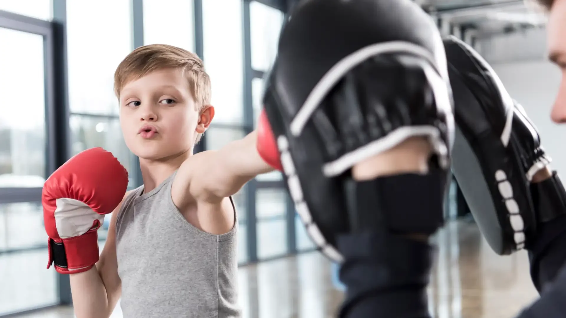 Garotinho treinando boxe com treinador em academia, focado na prática de esportes de combate.
