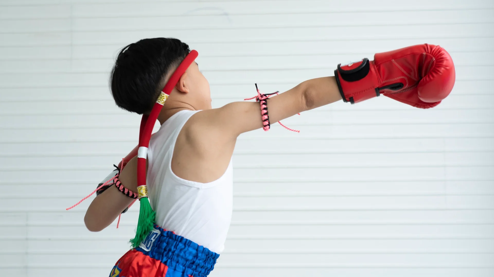 Menino pequeno treinando muay thai com luvas vermelhas, vestindo roupa de time e usando protetor de cabeça, demonstrando força e determinação.