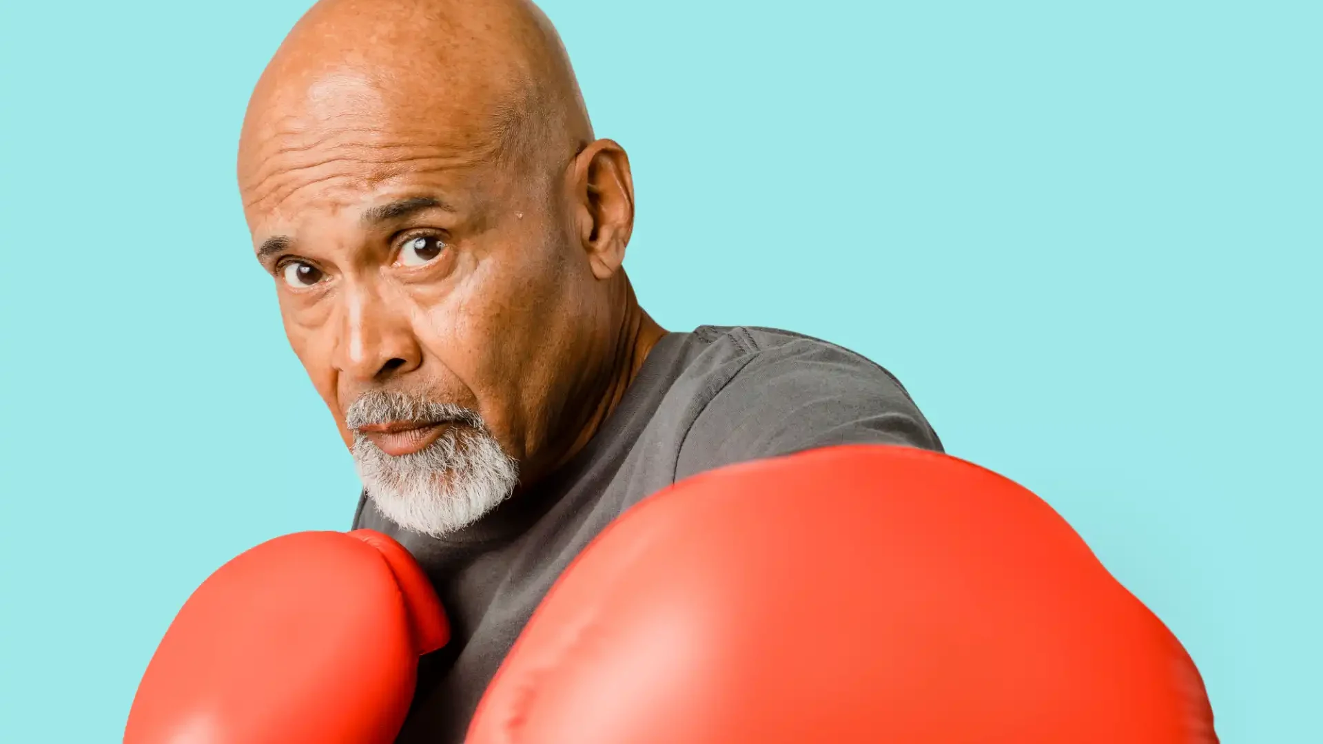 Homem mais velho com barba grisalha e careca, vestindo uma camiseta cinza escuro, usando luvas de boxe vermelhas, posando como um boxeador enérgico com fundo azul claro.