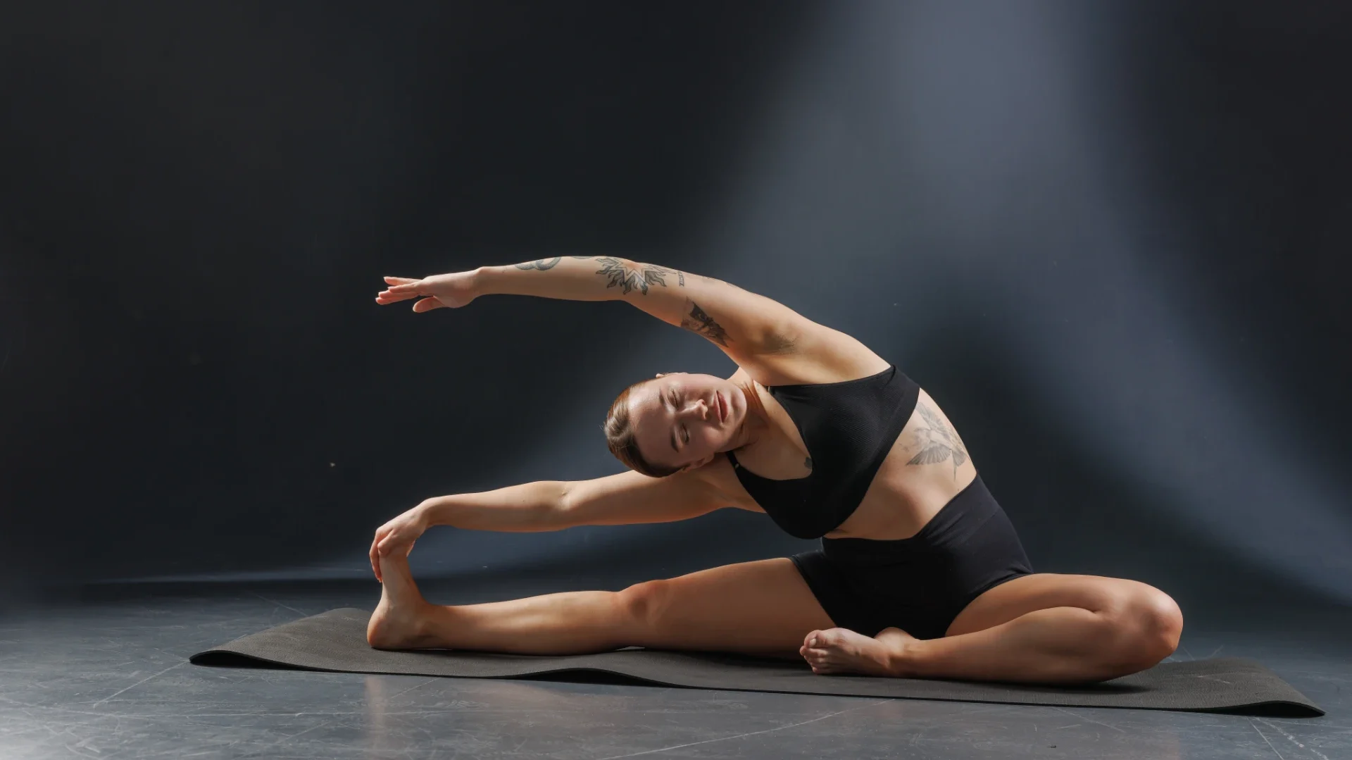 Mulher alongando em meio a um ambiente escuro, realizando um movimento sentado com um braço ao lado da cabeça, vestindo roupas de ginástica pretas.