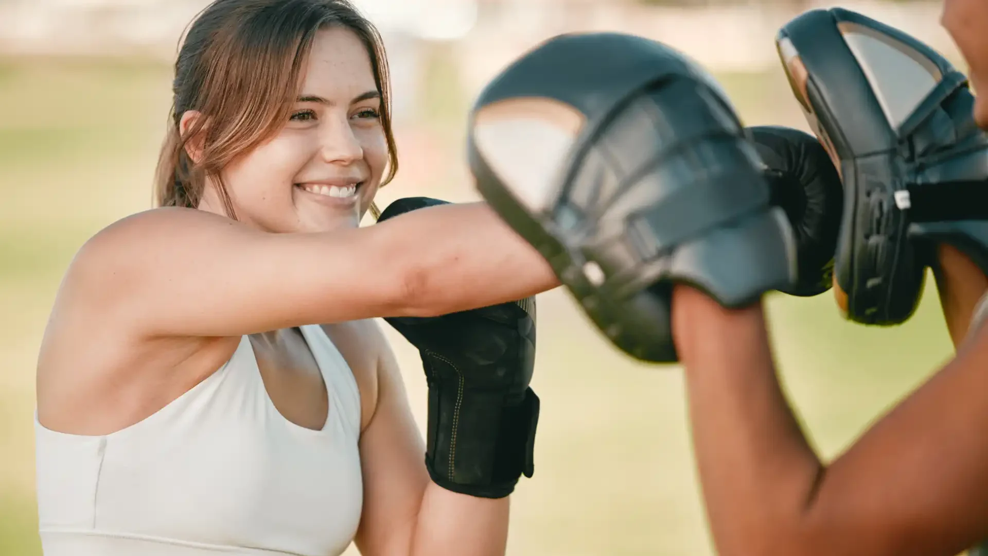 Mulher praticando boxe ao ar livre com um instrutor, sorridente e usando luvas de boxe, durante treino em parque ao ar livre.