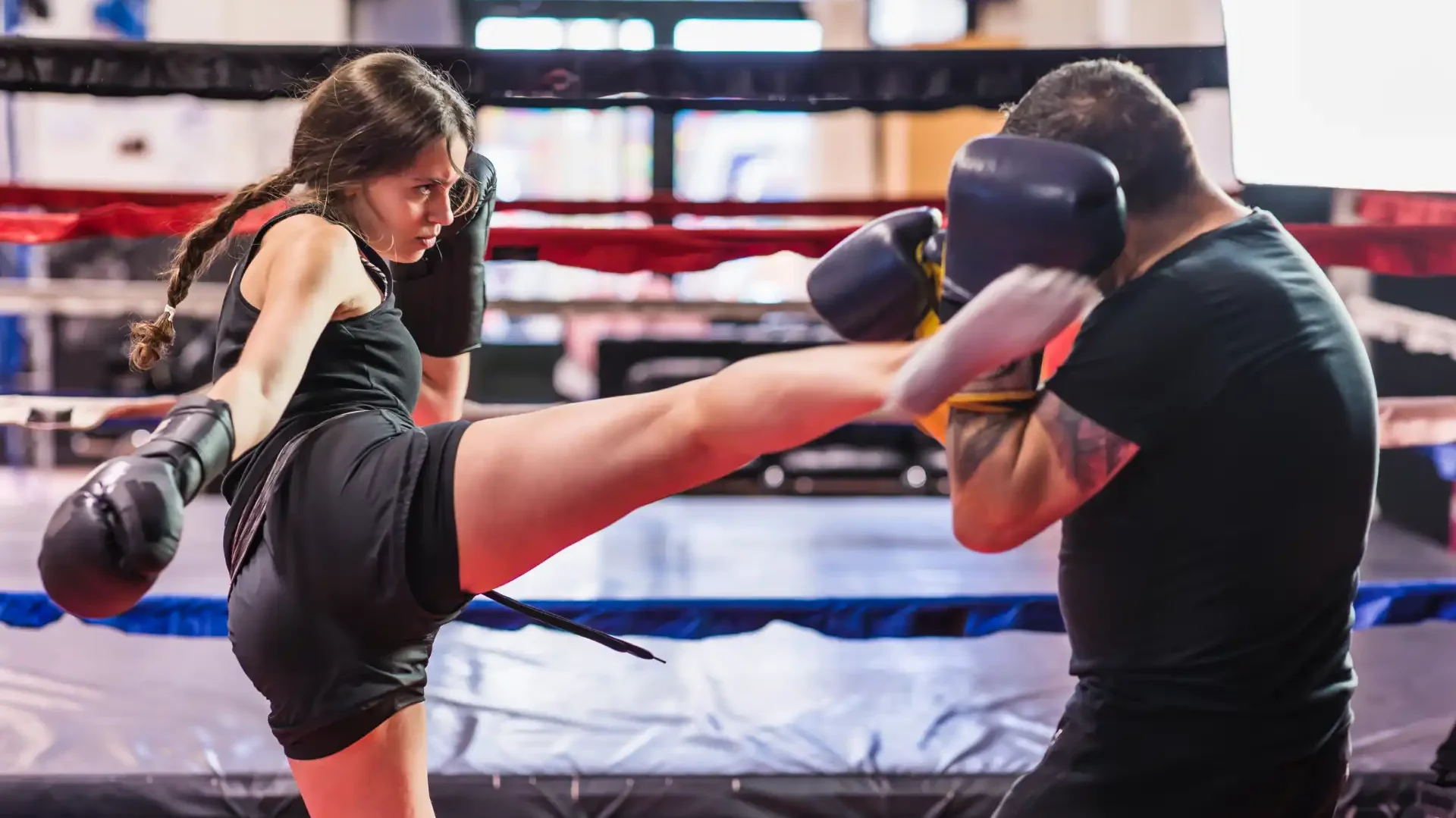 Mulher treinando muay thai na academia, realizando um chute em um treinador com luvas de boxe.