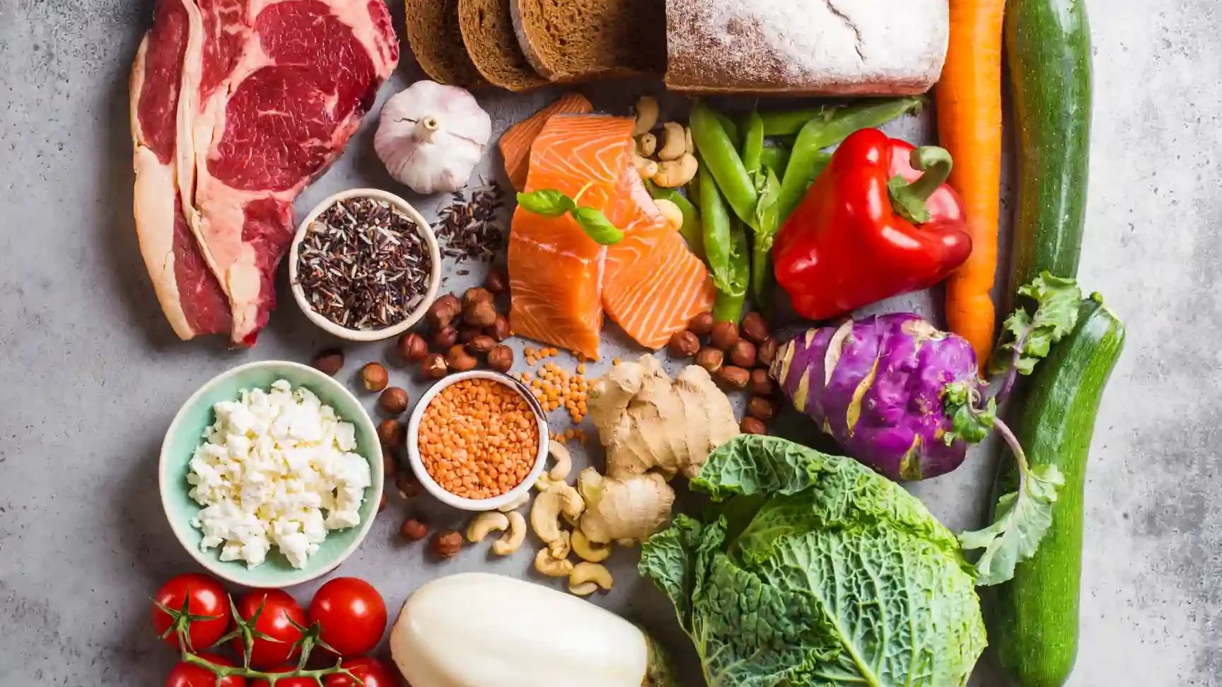 Alimentos variados e saudáveis para dieta equilibrada nutritiva incluindo carne, vegetais, grãos e temperos, dispostos sobre uma superfície de concreto.