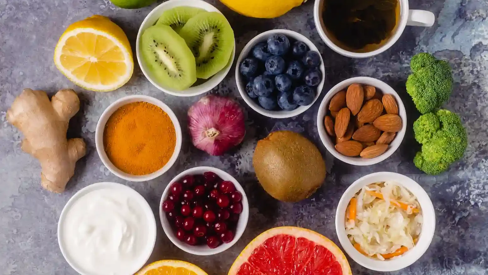 Variedade de frutas frescas e ingredientes saudáveis dispostos em uma superfície, incluindo limão, morango, kiwi, mirtilo, gengibre, castanhas, iogurte e chá.