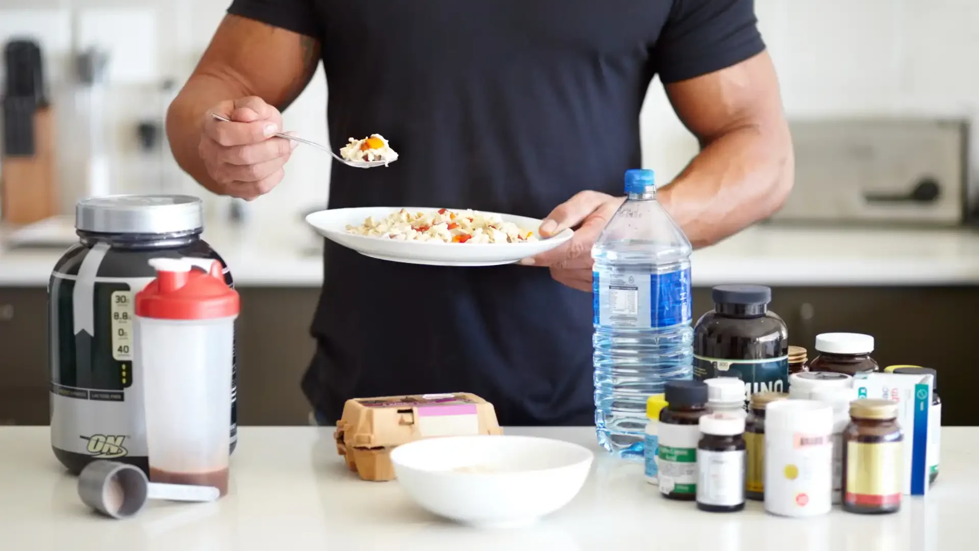 Homem em uma cozinha com diversas vitaminas e suplementos, segurando um prato de comida saudável, promovendo uma alimentação balanceada.