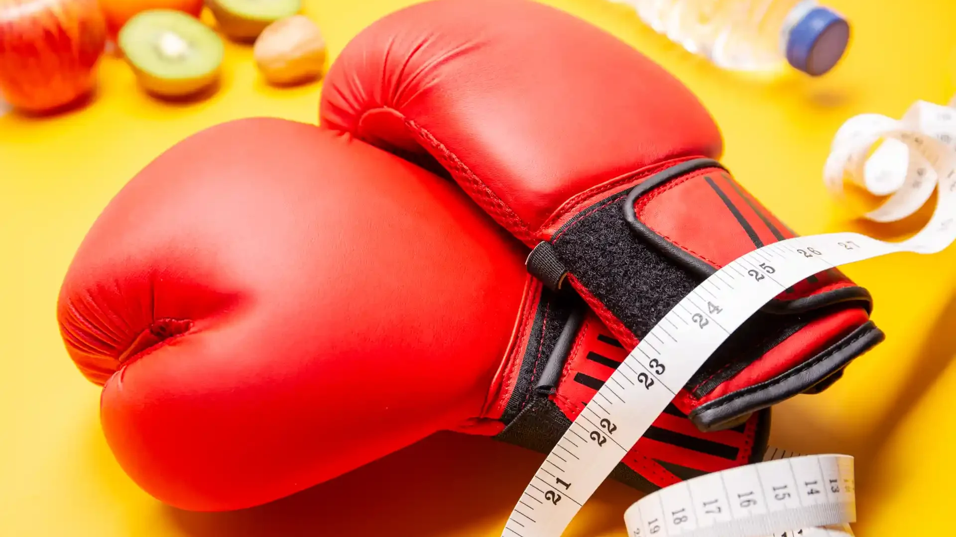 Luvas de boxe vermelhas com fita métrica ao redor, sobre fundo amarelo, símbolos de saúde e fitness ao fundo.