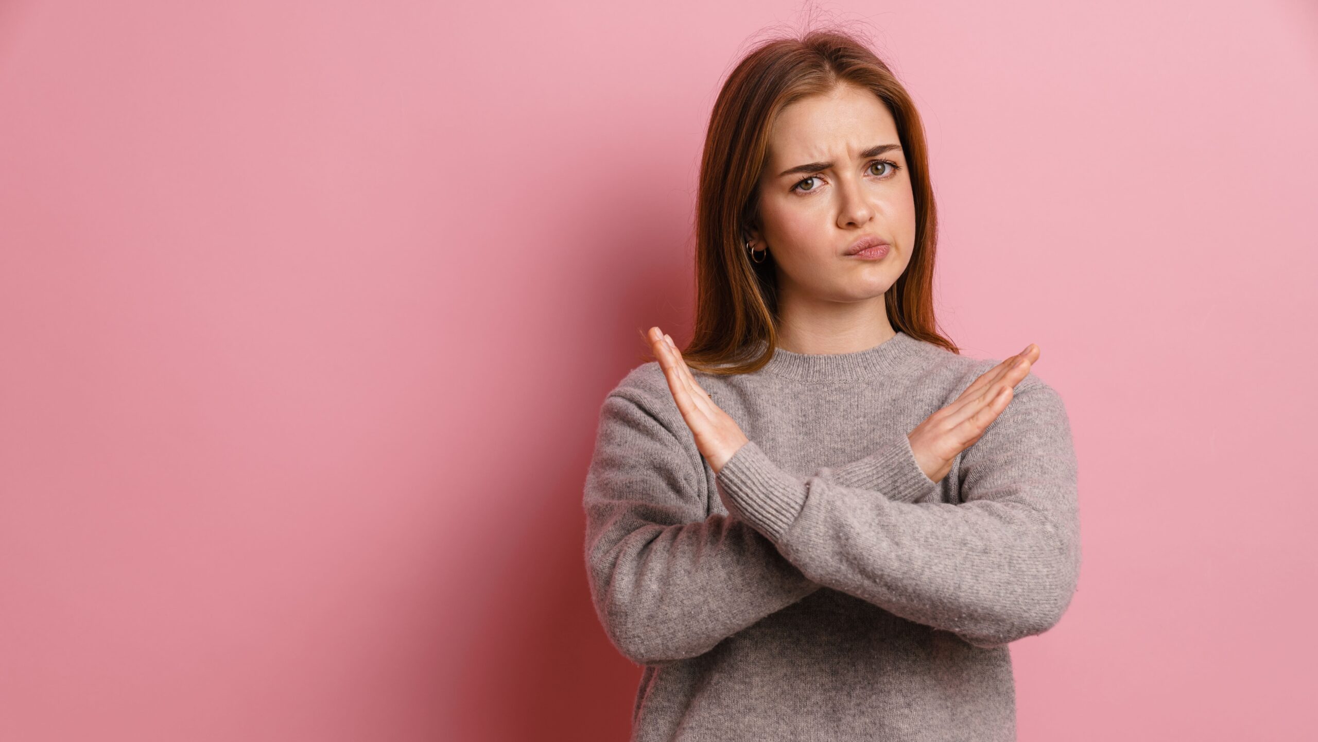 Mulher jovem com expressão de rejeição ou recusa diante de fundo rosa, usando suéter cinza, demonstrando discordância.