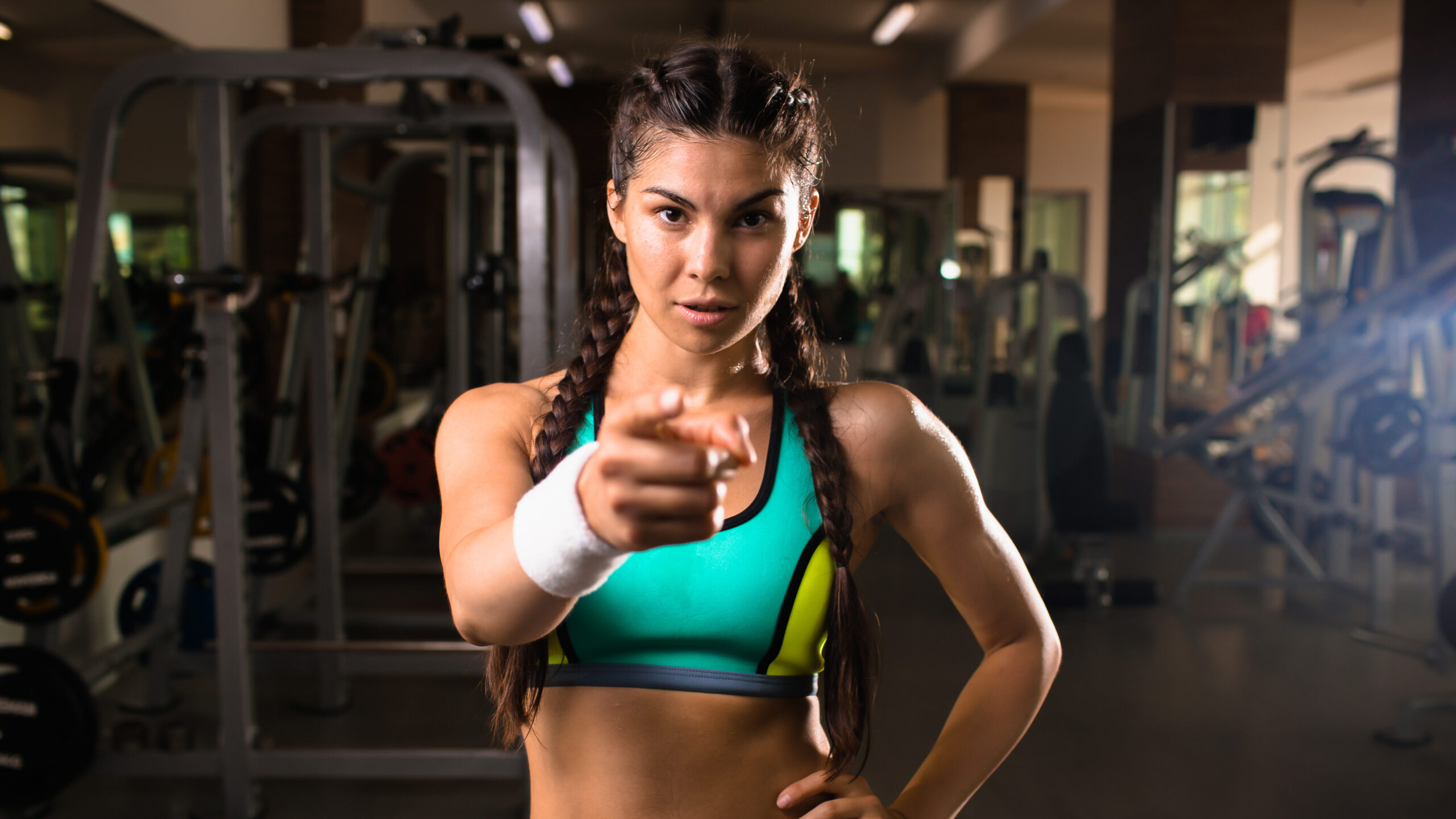 Mulher atleta no ginásio apontando diretamente para a câmera, vestindo roupa esportiva, com bom preparo físico e foco em treinamento de alta intensidade.