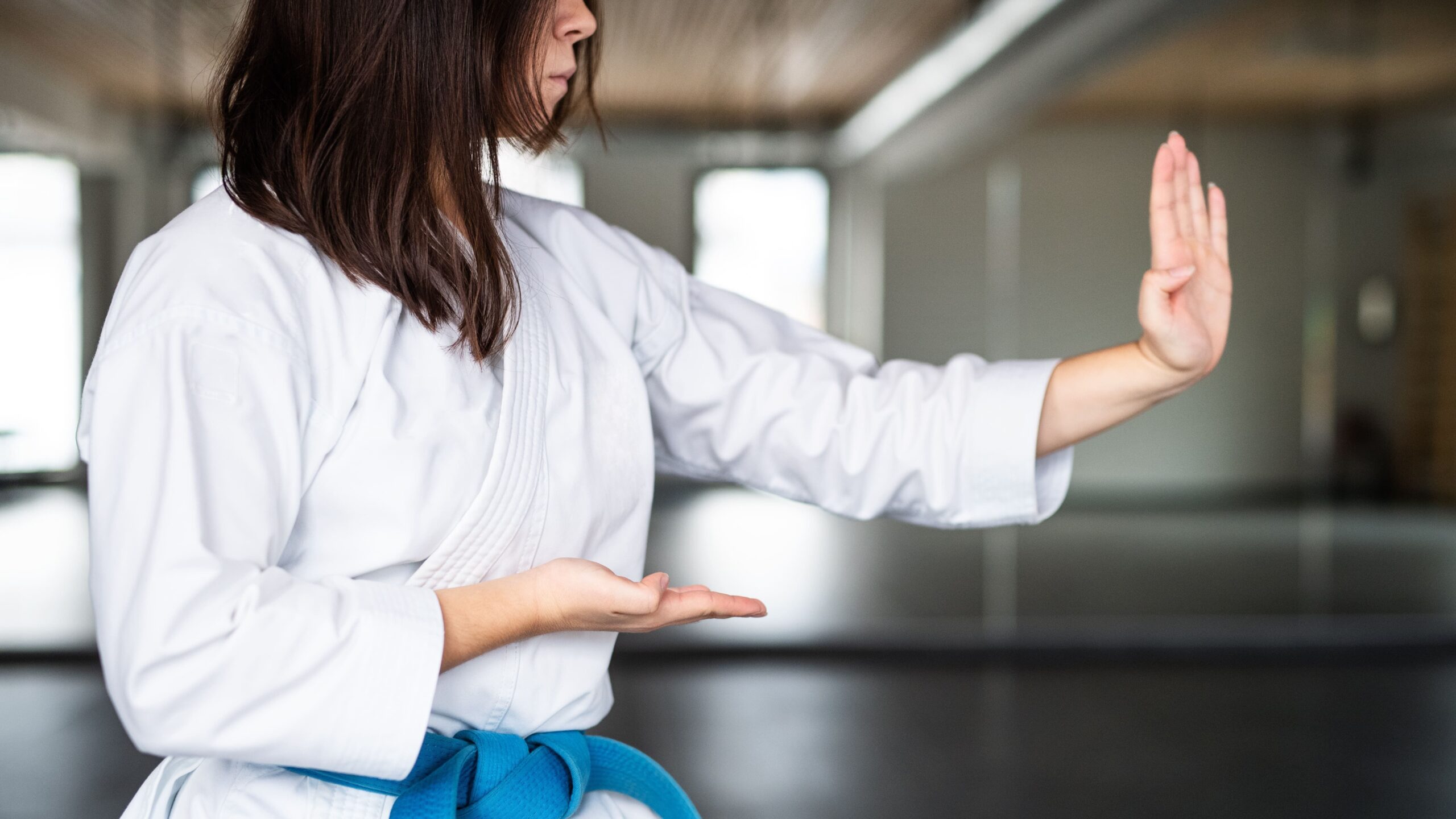 Instrutora de lutas demonstrando técnica de defesa em ambiente de dojo, com kimono branco e faixa azul, focada na prática de artes marciais.