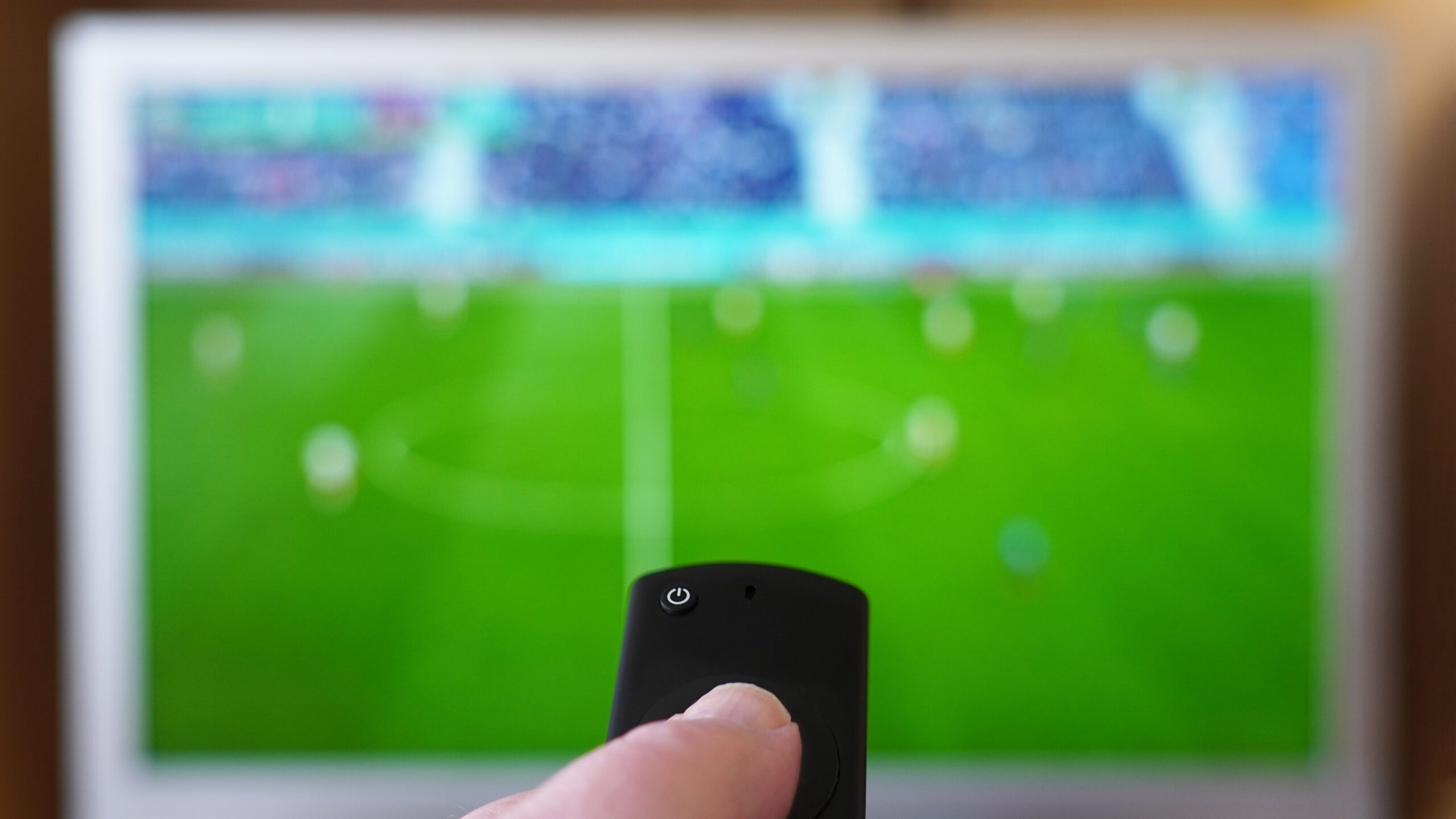 Pessoa assistindo a uma partida de futebol na televisão enquanto usa um controle remoto na mão, com uma imagem de estádio ao fundo.