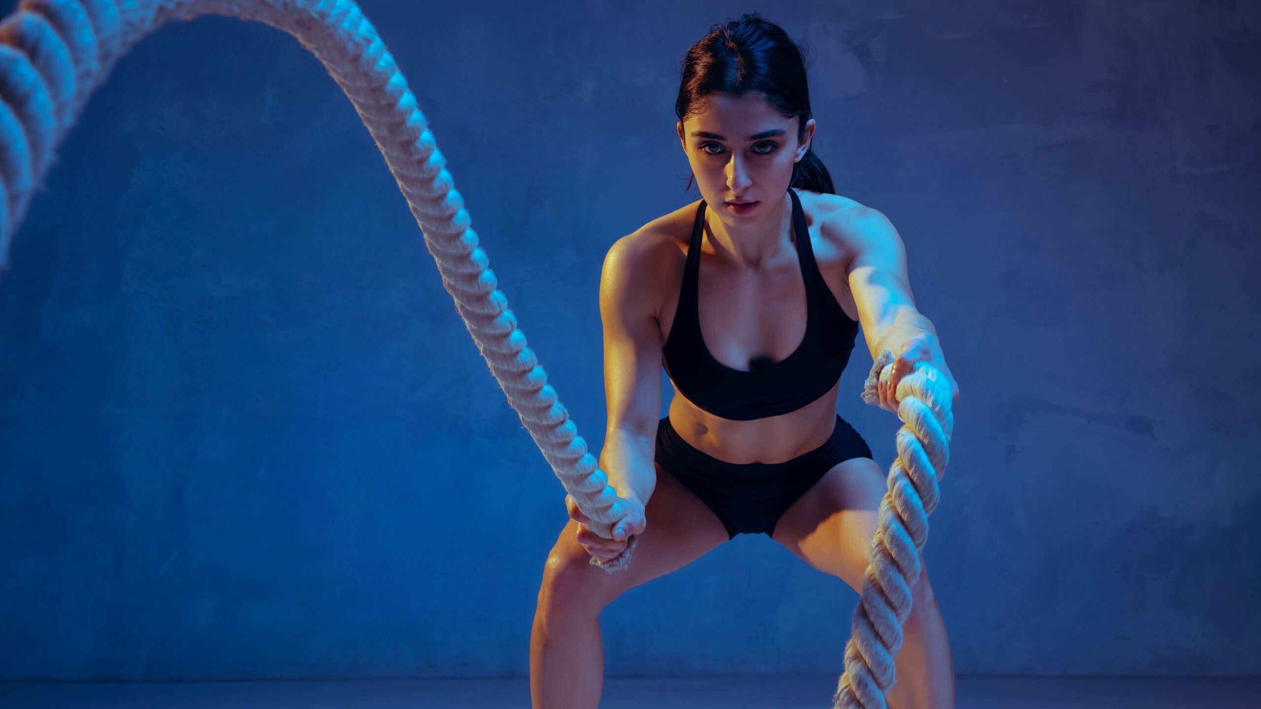 Mulher fitness treinando com cordas de batalha em ambiente de academia, vestindo roupa de ginástica preta, com foco em exercícios de força e resistência.