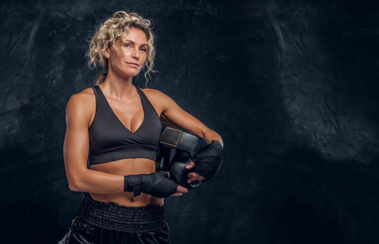 Mulher atleta com cabelos cacheados curtos, vestindo roupa de ginástica preta e luvas de boxe, segurando o protetor bucal na mão, pronta para treino de boxe.