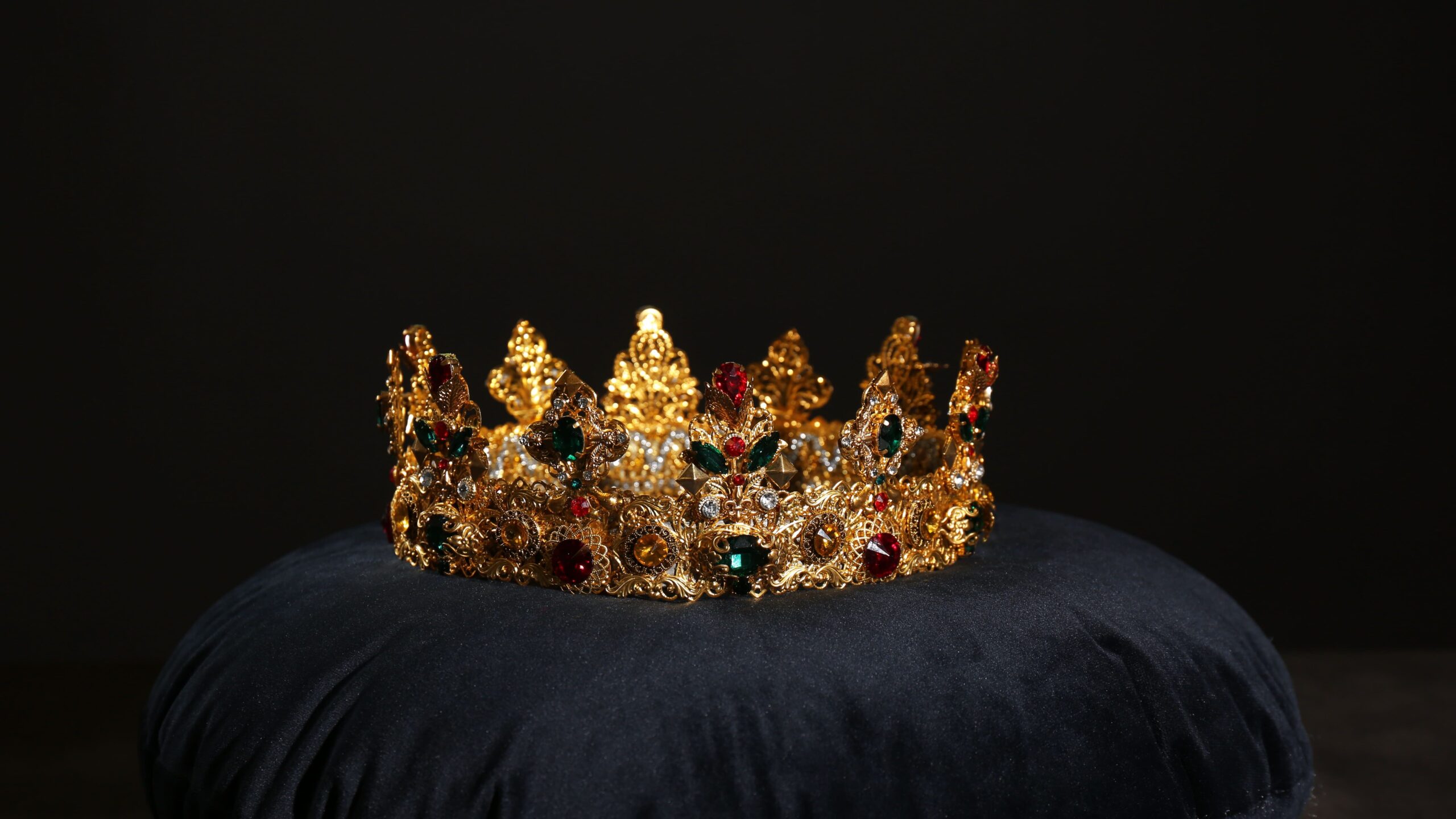 Imagem de uma coroa dourada decorada com pedras preciosas coloridas, exalada de luxo e riqueza, colocada sobre uma almofada preta ao fundo preto.