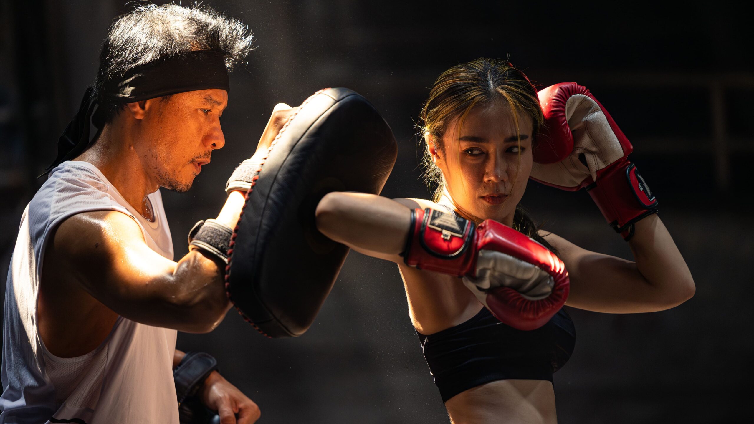 Mulher treinando boxe com técnico, golpes com luvas vermelhas, ambiente de academia de boxing.