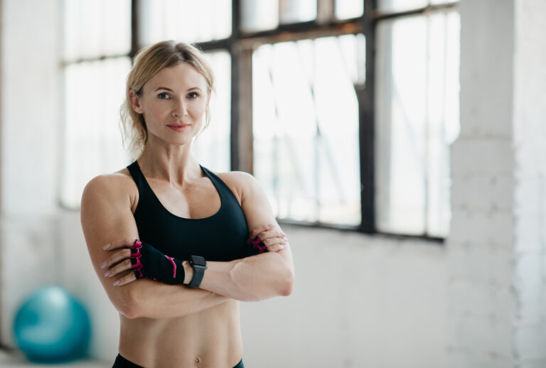Mulher treinando força física em ginásio com expressão confiante, vestida com roupas de esporte pretas e luvas de treino, ambientes de academia ao fundo.