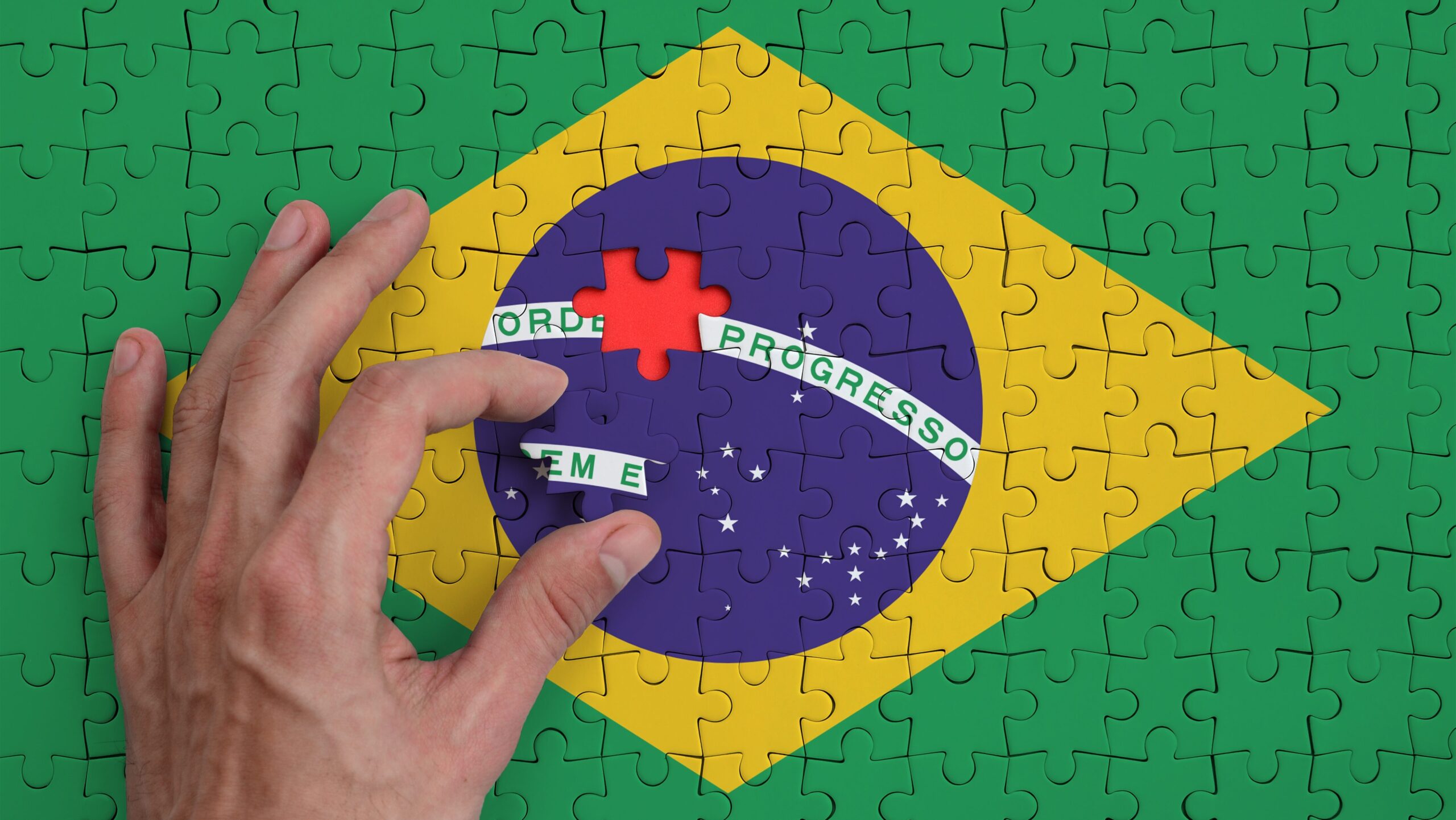 Quebra-cabeça de uma bandeira do Brasil sendo montado, com uma mão ajustando uma peça vermelha na cena.