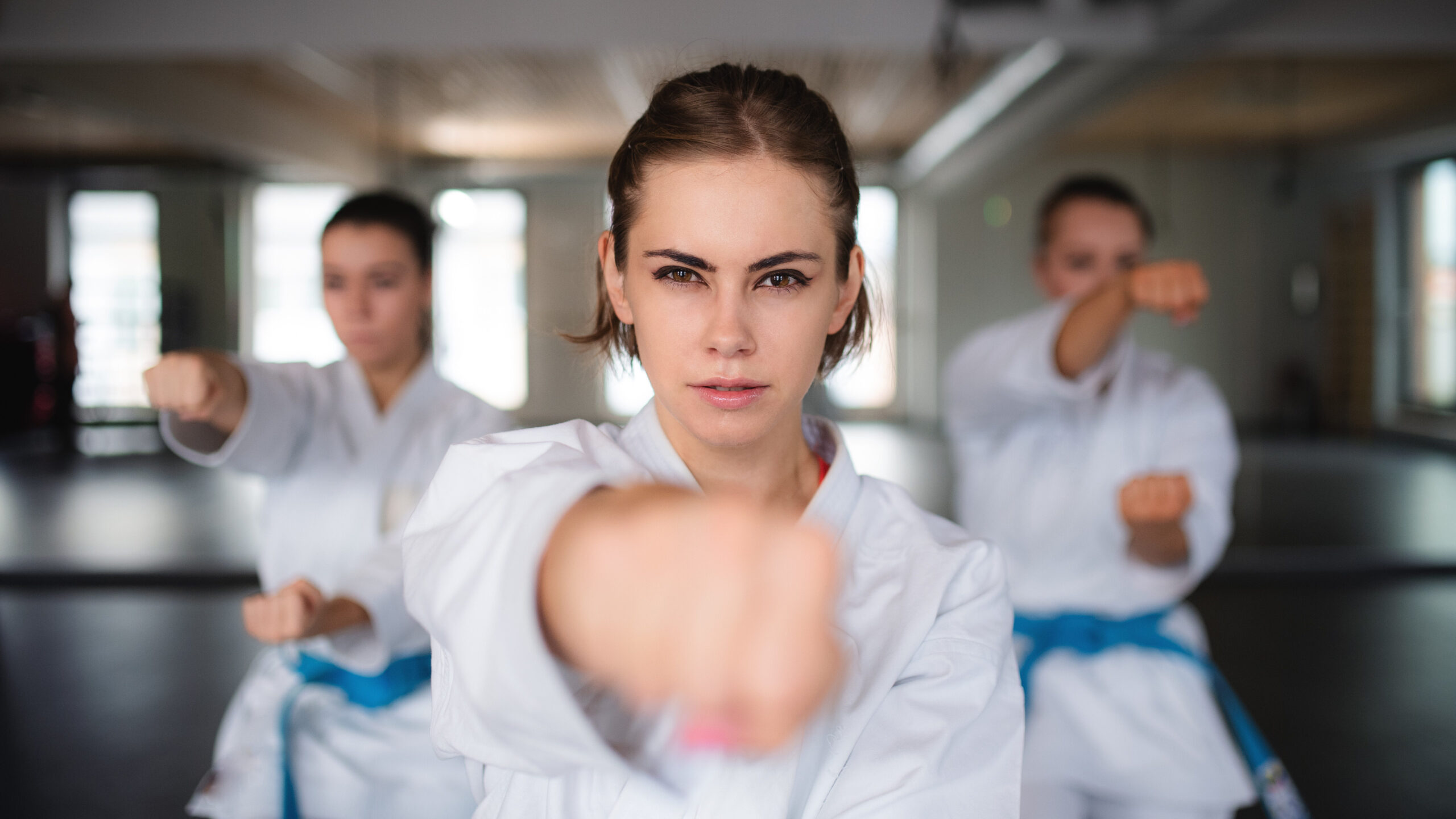 Três mulheres praticando arte marcial em uma academia, focadas em uma técnica de defesa pessoal, demonstrando força, disciplina e determinação.