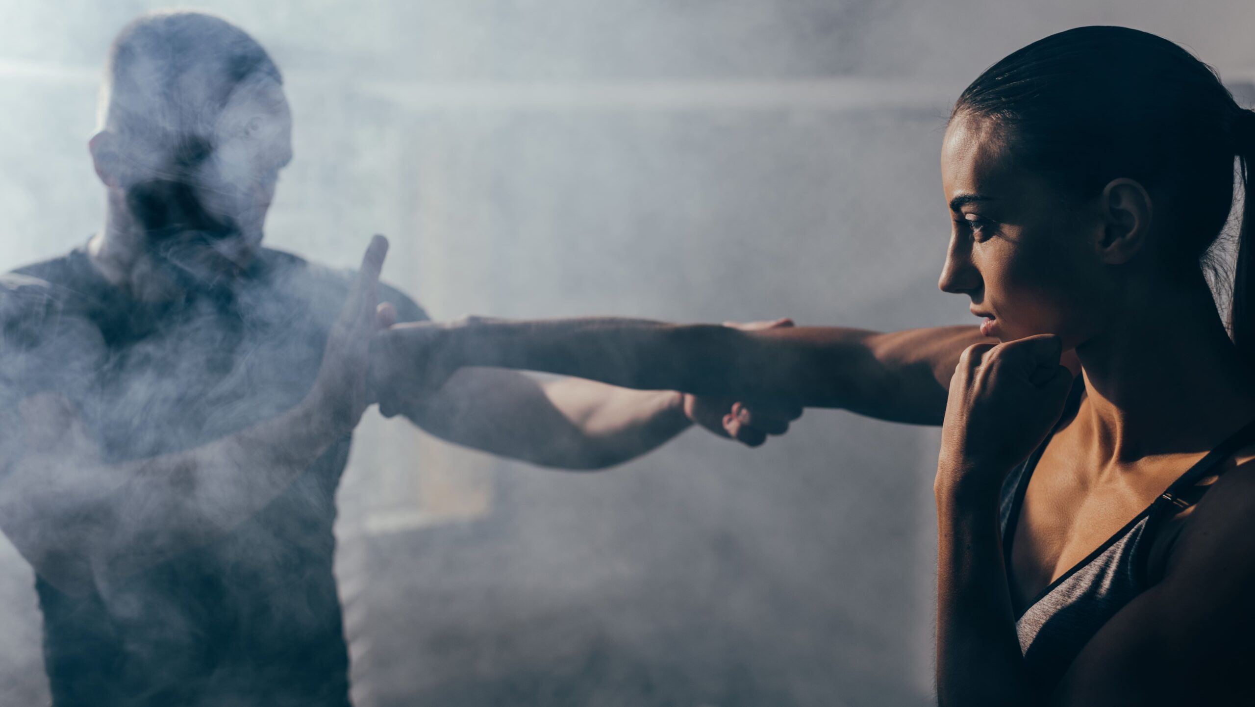 Mulher treinando luta com homem, ambos com postura de artes marciais e expressões de concentração, em ambiente de treino com pouca luz