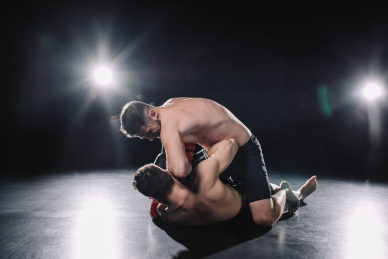Lutadores de MMA em uma luta intensamente travada, com um lutador dominando o outro no chão, sob iluminação dramática destacando a ação no ringue.