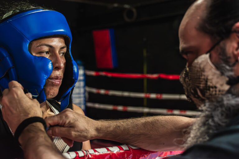 Treinadora ajudando uma lutadora de boxe a ajustar seu capacete antes de uma luta, em uma academia de boxe, mostrando preparação e técnica esportiva.