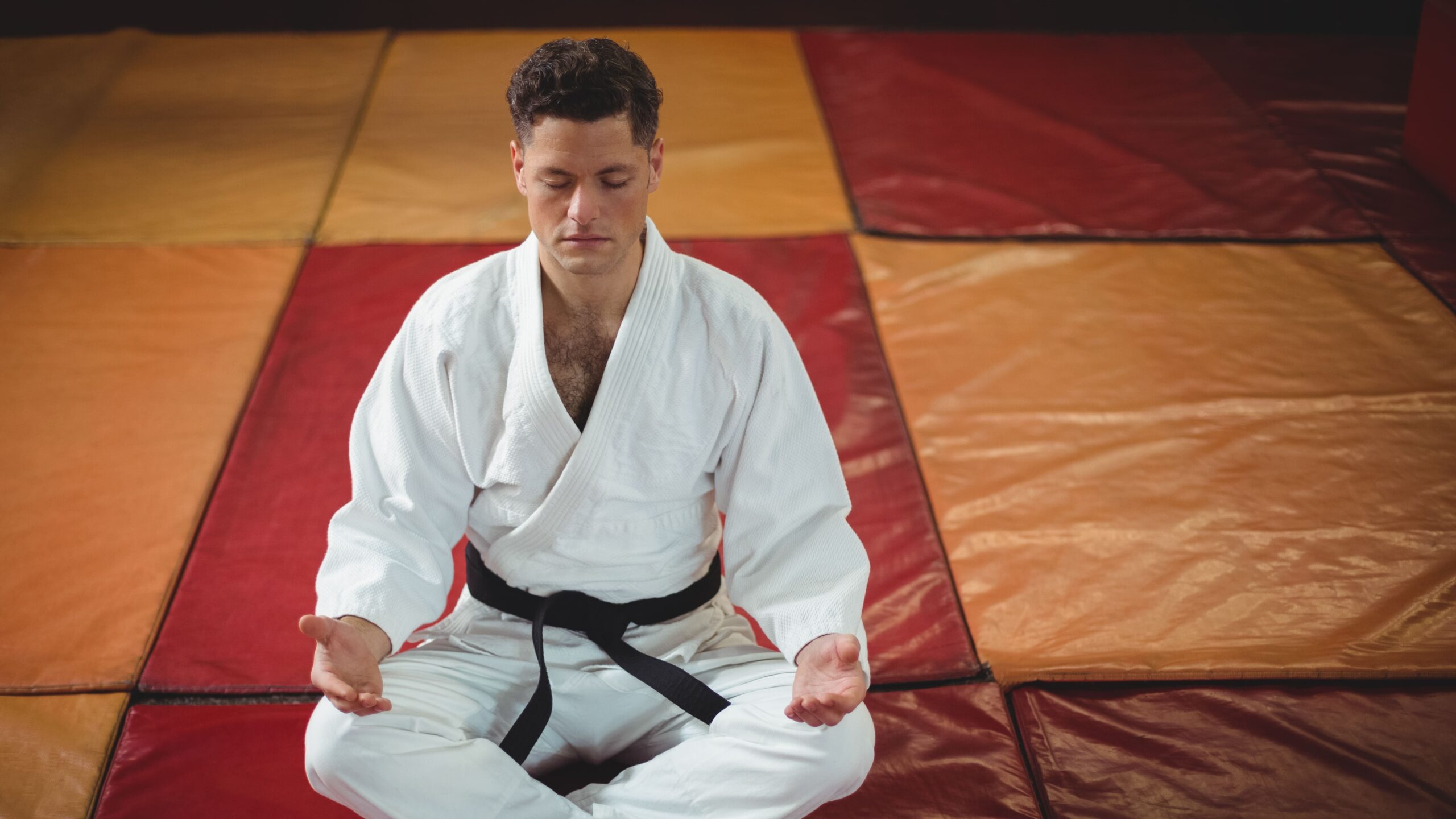 Homem praticando meditação em uma academia de judô, sentado em posição de lótus com olhos fechados, vestindo kimono branco e em um ambiente com tatames coloridos.