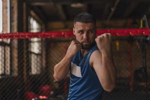 Homem lutando em um ringue de boxe, com tronco à mostra e colete azul, em postura de guarda, apertando os punhos e olhando focado para o oponente.