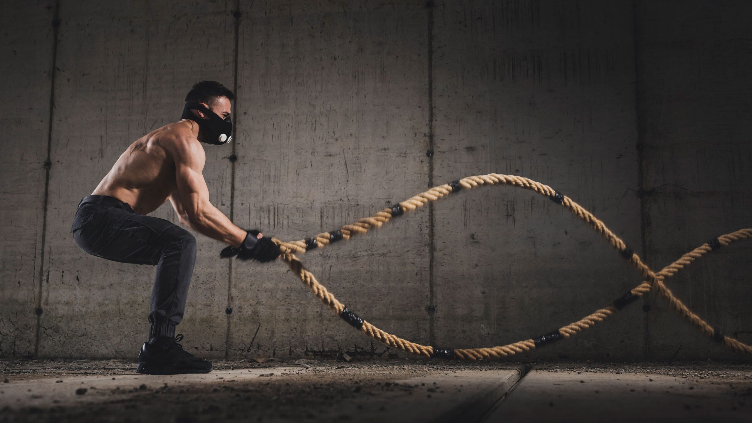 Homem agachado em treino funcional com cordas navais, puxando as cordas em movimento repetitivo em um ambiente industrial com parede de concreto