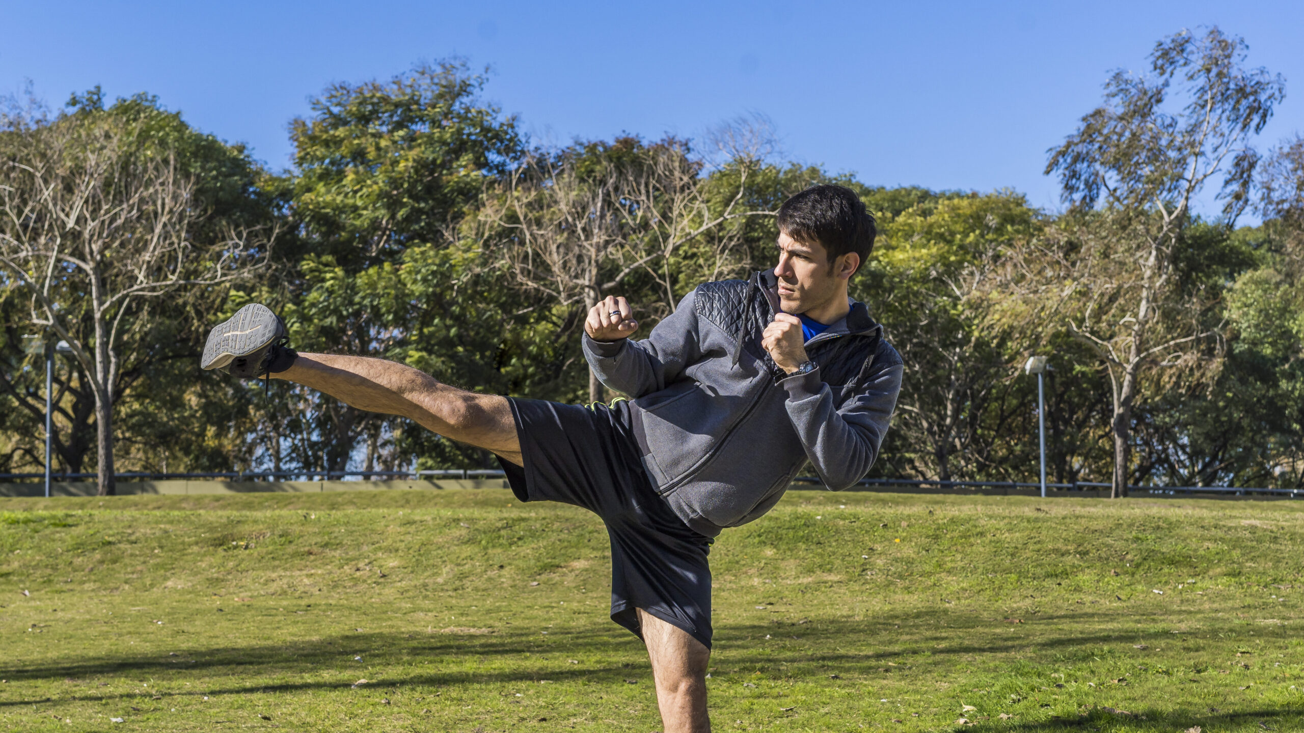 Homem em parque fazendo movimento de chute com árvores e céu azul ao fundo