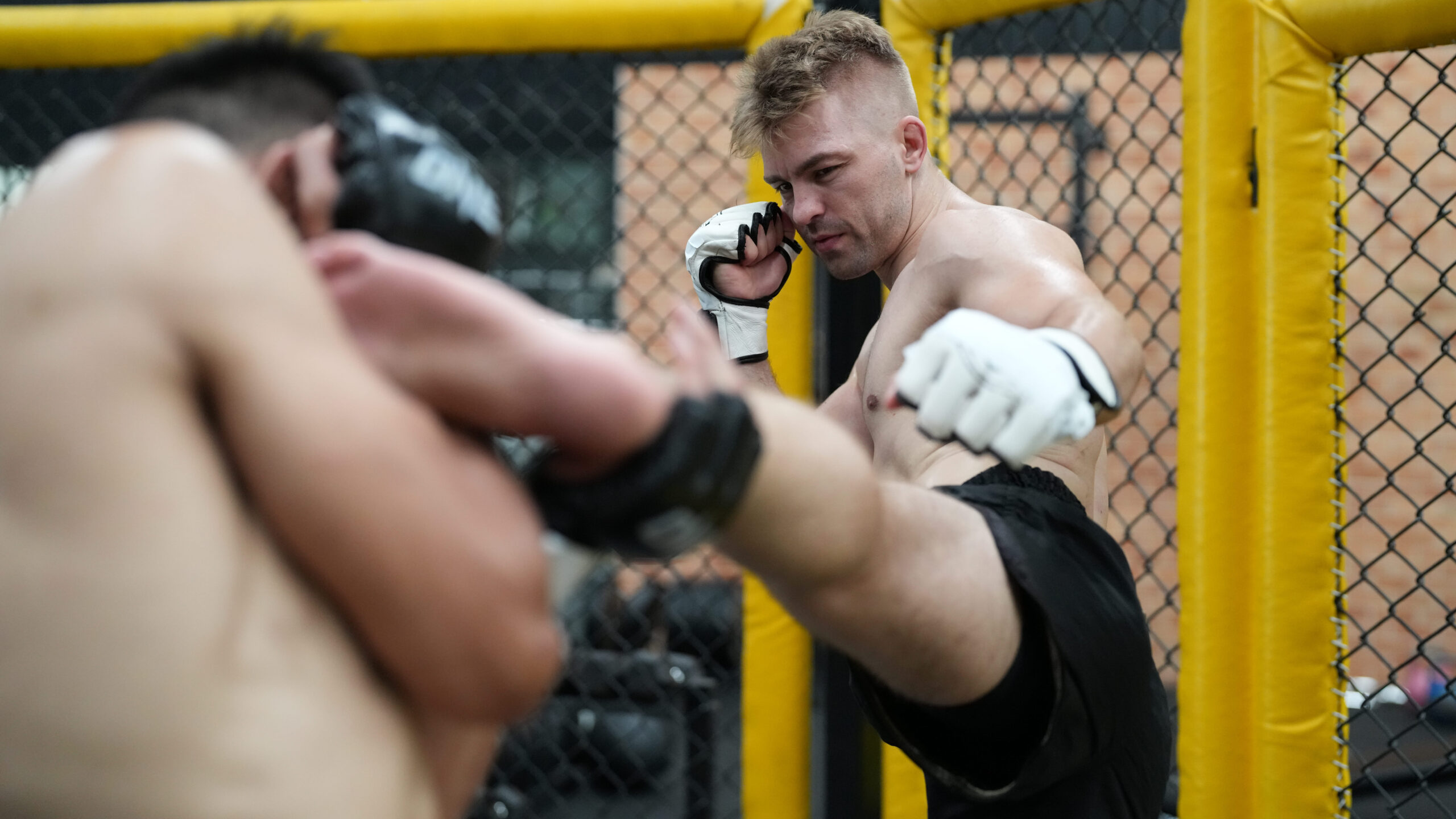 Lutador de MMA em uma luta dentro do octógono, com luvas e postura de combate, enquanto o adversário aparece em primeiro plano desfocado.