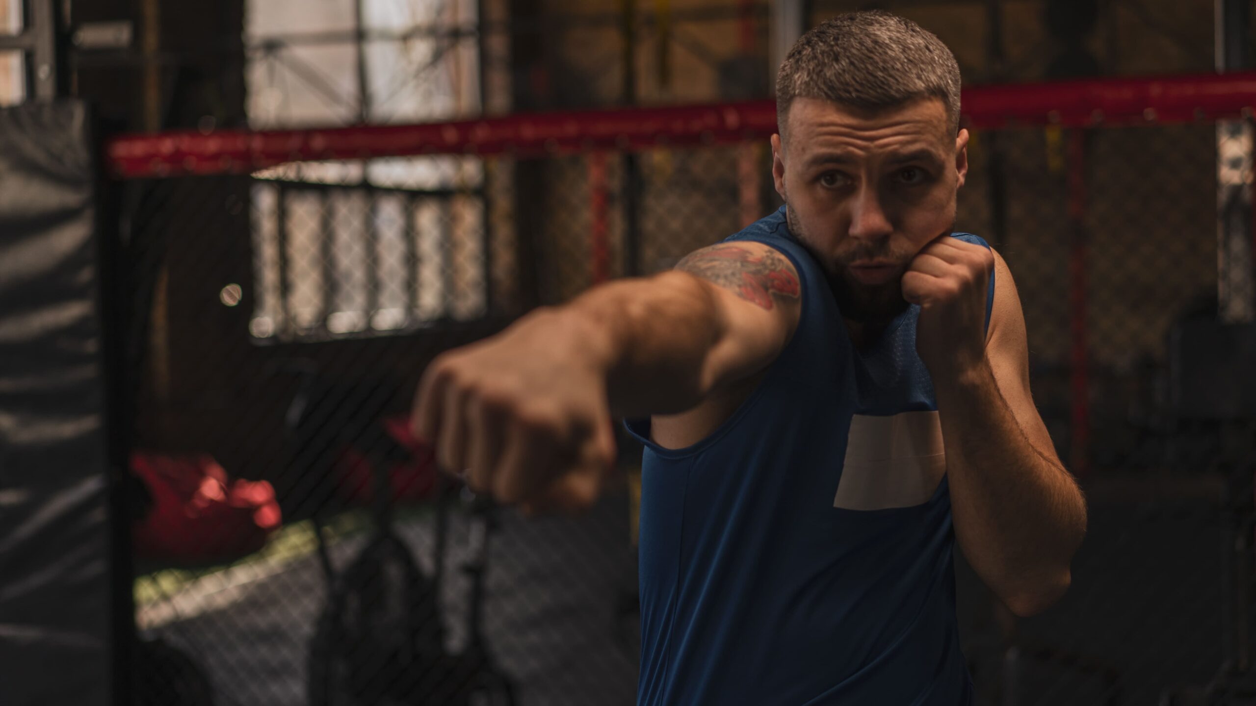 Lutador de boxe em treino dentro do ringue, com o braço estendido e pose de ataque, usando camisa azul e encarando o oponente em um ambiente de arena com grades e iluminação de luta.