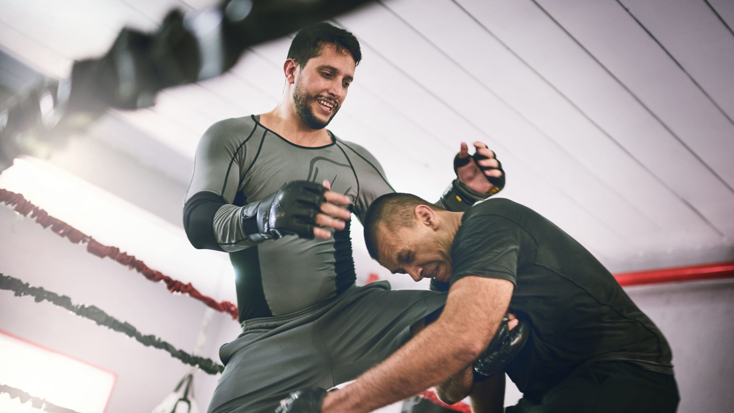 Dois homens treinando boxe em uma academia, com luvas e postura de golpe, um deles aplica movimento enquanto o outro se prepara para defender, em ambiente de treino com cordas de boxe.