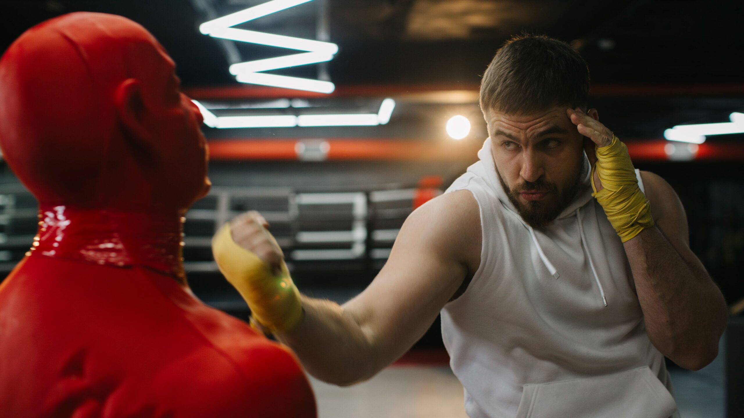 Dois boxeadores em um ringue, com um lutador de camisa branca e luvas amarelas se protegendo e o outro em uniforme vermelho avançando durante uma luta de boxe em ambiente de treino com iluminação de ginásio.