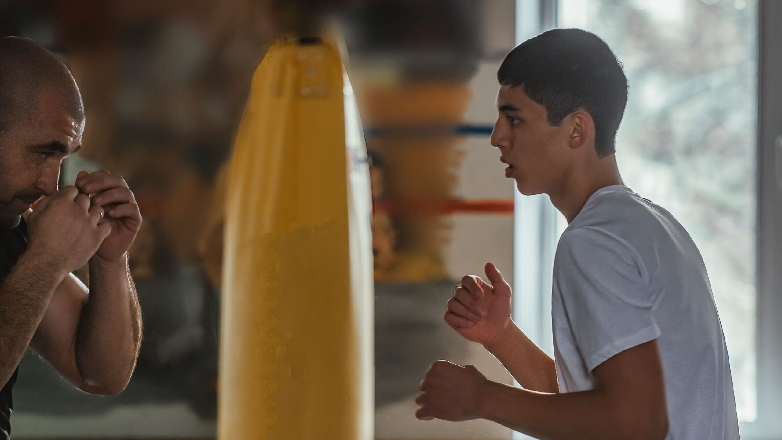 Homem em sala de treino ajustando a respiração e observando o companheiro ao lado de um saco de pancadas, com foco e postura de preparação para treino de boxe