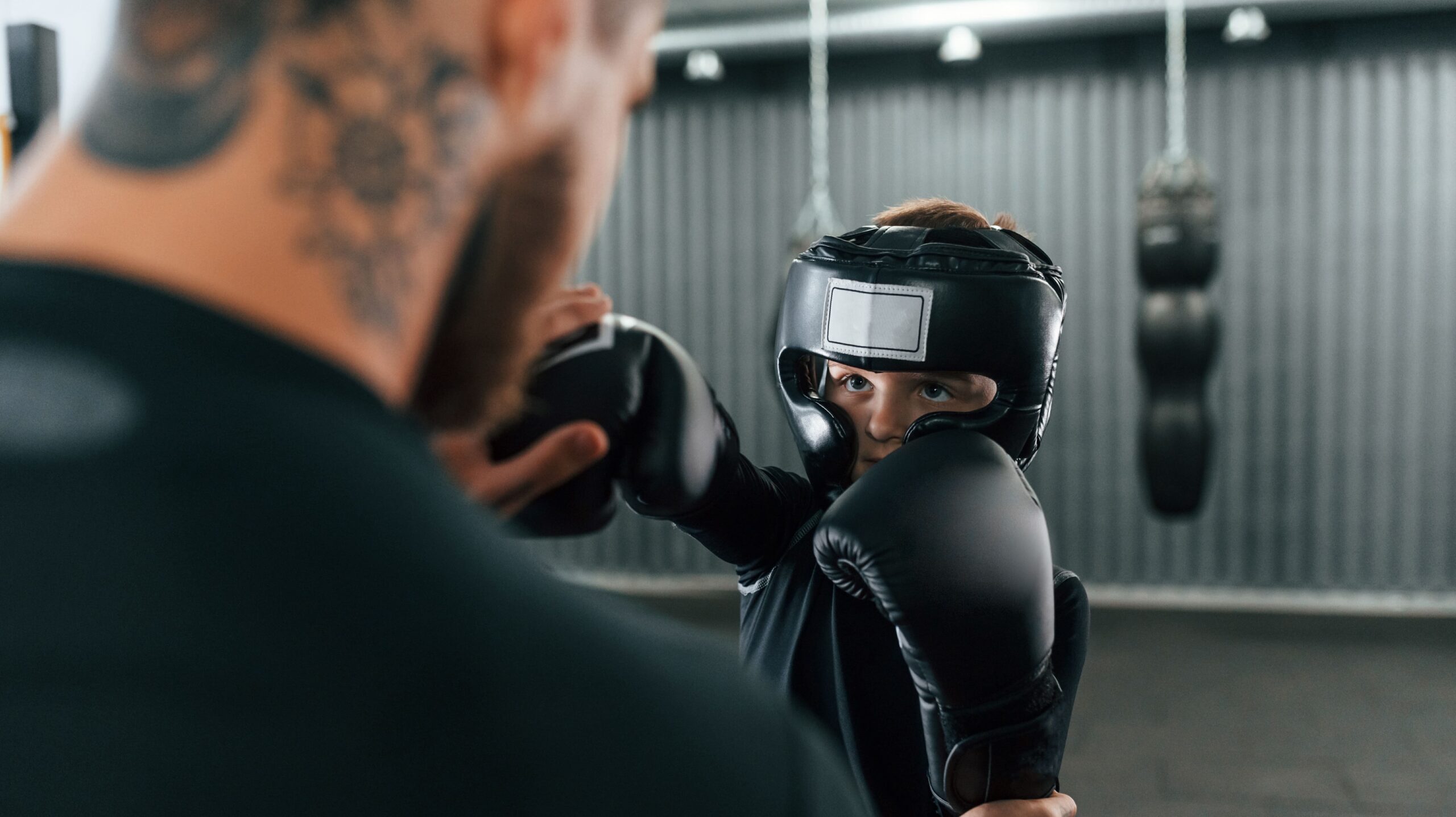Treino de boxe em academia: lutadora jovem com capacete e luvas negras ajusta a guarda e encarando o parceiro, que usa equipamentos de proteção, em ambiente com saco de pancadas ao fundo.