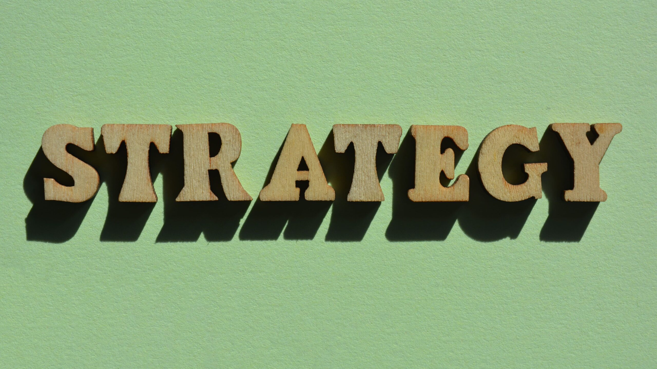 Texto “STRATEGY” em letras 3D estilo recorte, com preenchimento escuro e acabamento em tom de madeira, sobre fundo verde claro, criando contraste e efeito de profundidade.