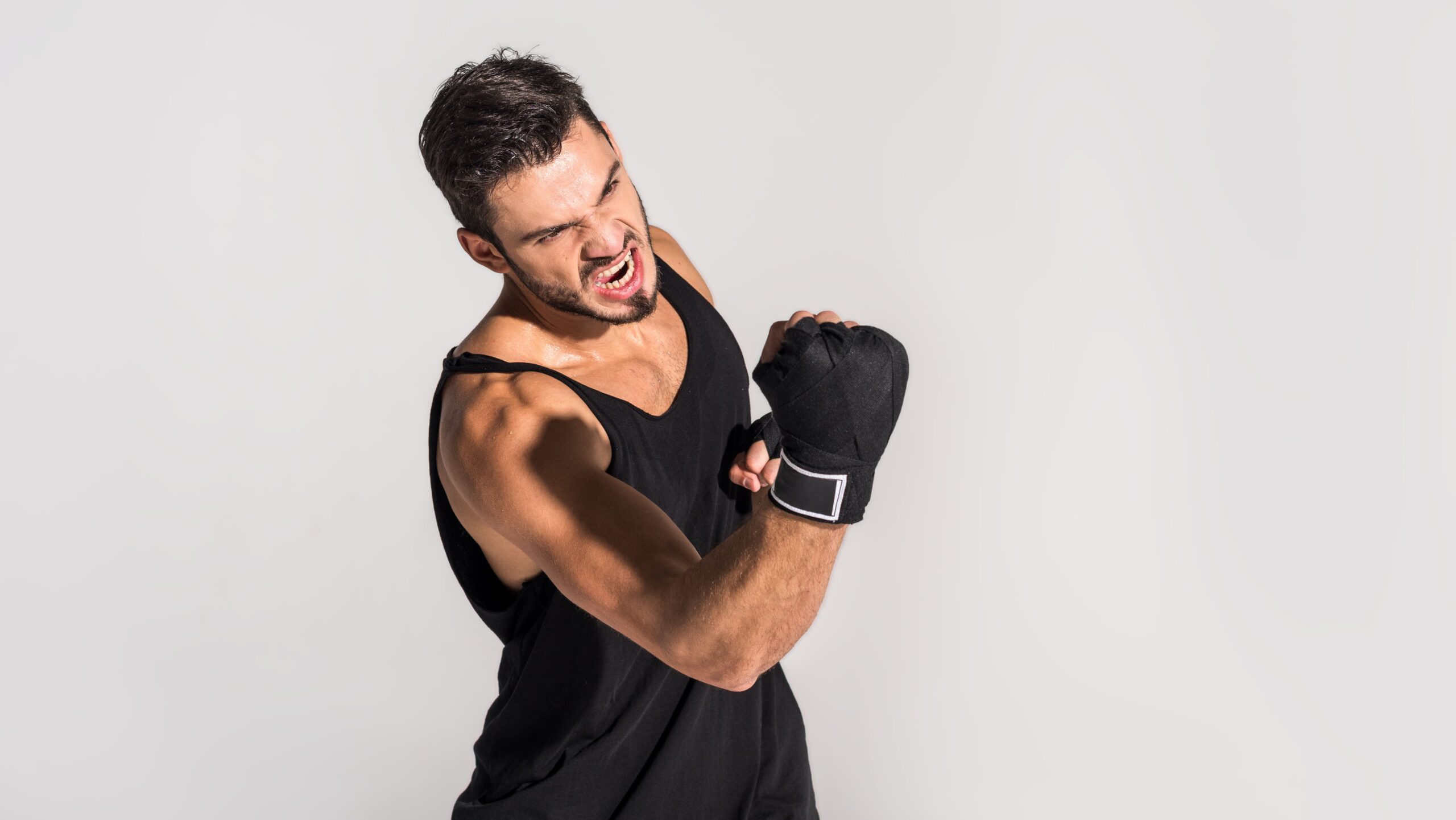 Homem jovem com camiseta preta em pose de luta, segurando uma luva de treino e com expressão focada, contra fundo branco, sugerindo treino de boxe ou artes marciais.