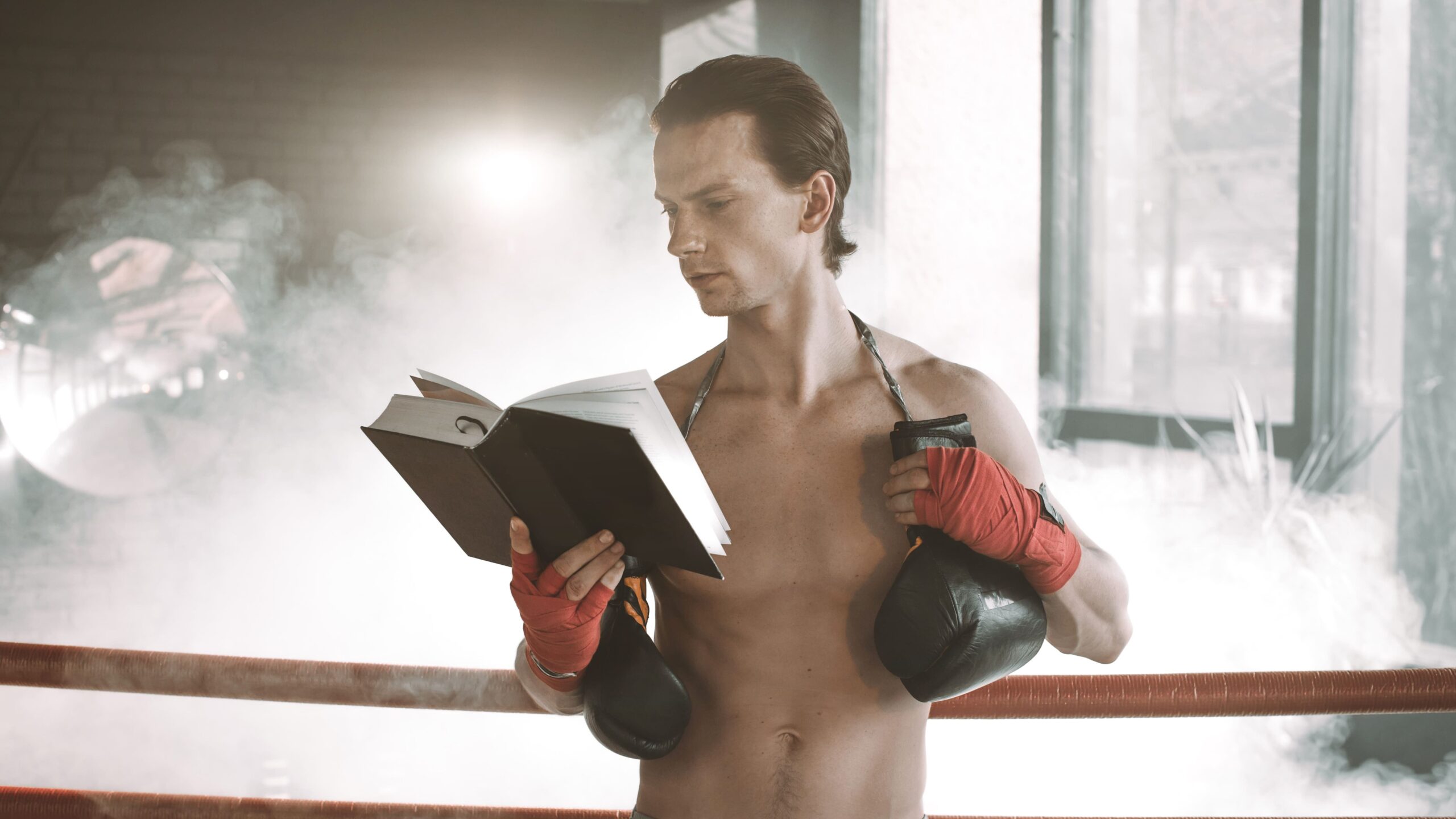Homem atleta em ambiente de academia, lendo um livro aberto enquanto segura luvas de boxe, diante de janelas com luz natural e fumaça suave ao fundo.