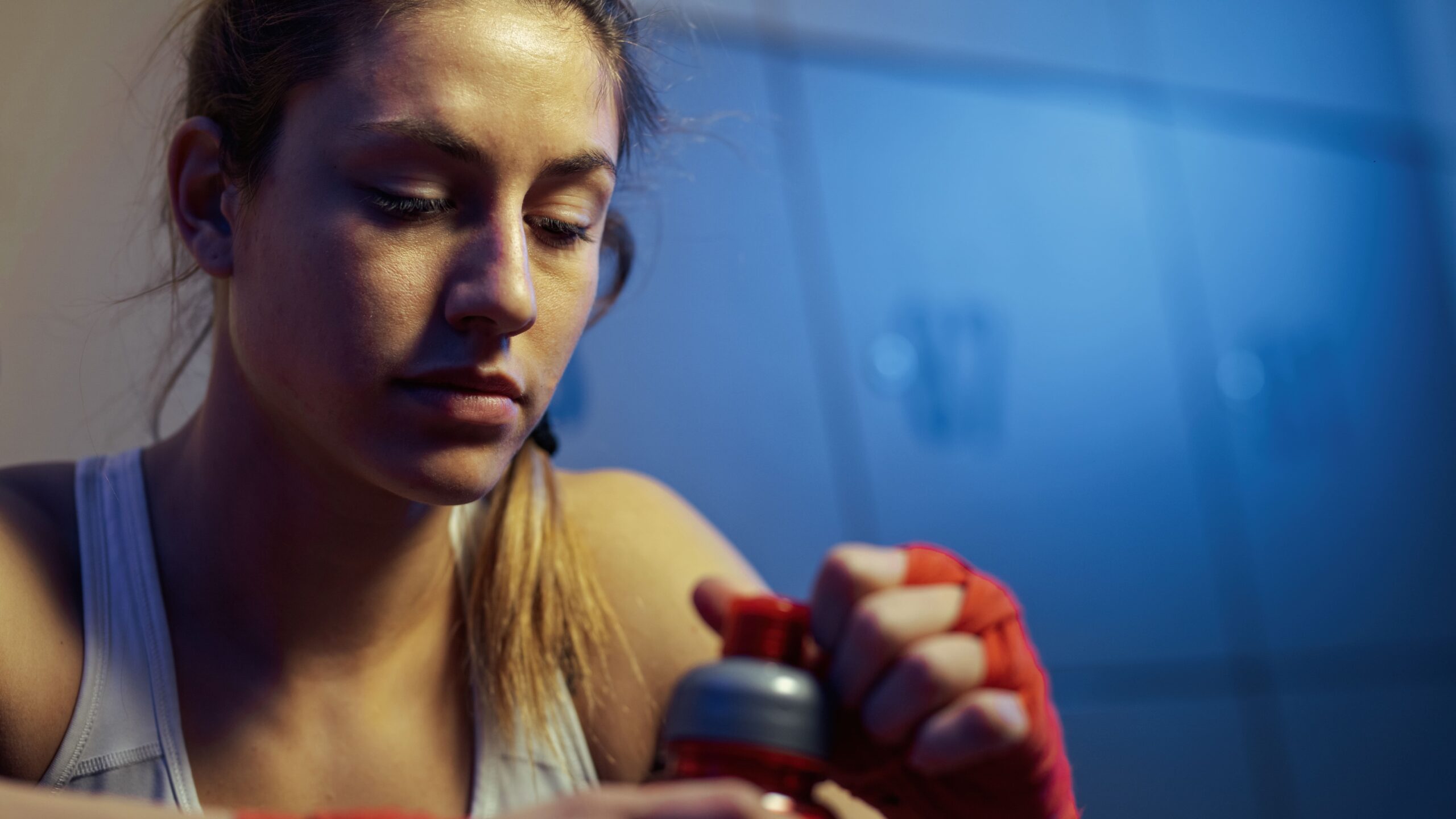 Atleta em primeiro plano, usando luvas de proteção, segurando um frasco tipo spray durante treinamento em ambiente esportivo, com luz azul ao fundo.