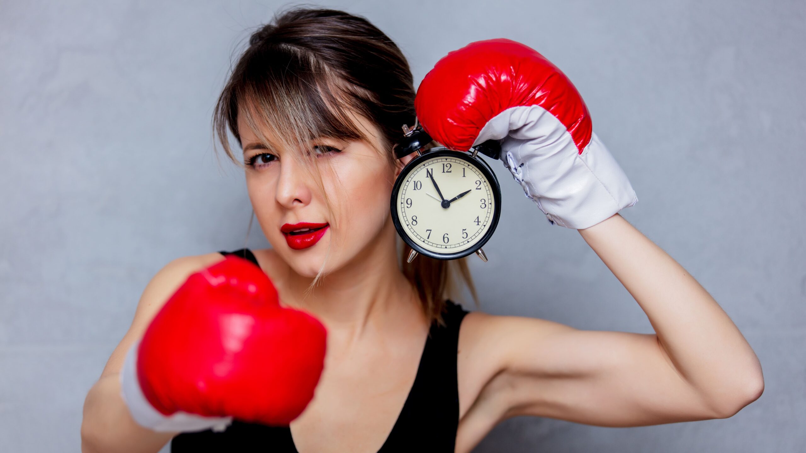 Mulher em postura de luta usando luvas de boxe vermelhas, segurando um relógio de despertador preto como símbolo de tempo, com fundo neutro claro.