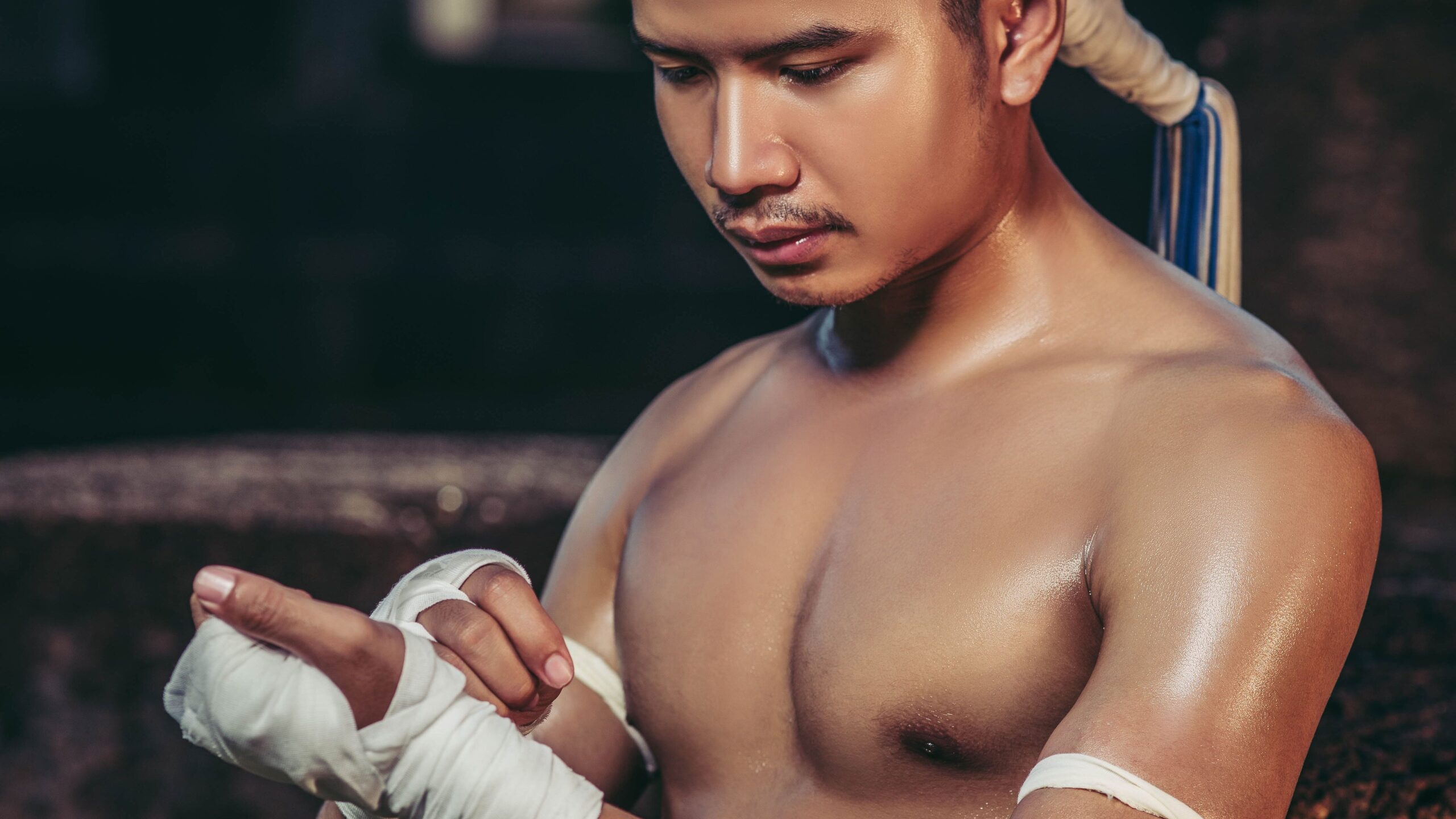 Lutador de Muay Thai em traje tradicional ajustando a bandagem branca na mão, com mongkhon na cabeça e kruang no braço.