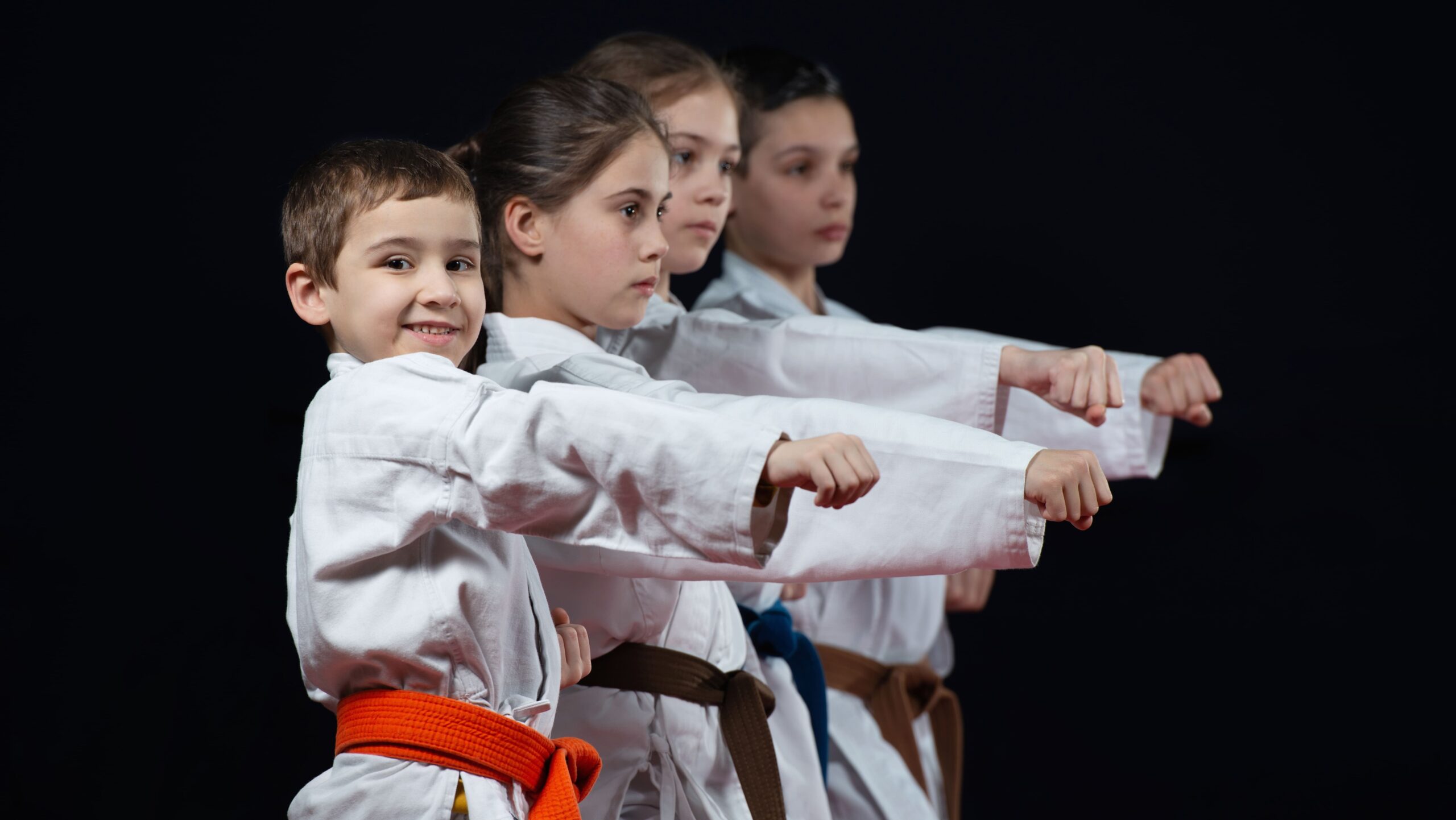 Quatro crianças de diferentes idades em fila vestindo quimonos, executando juntas o movimento de soco durante treino de artes marciais.
