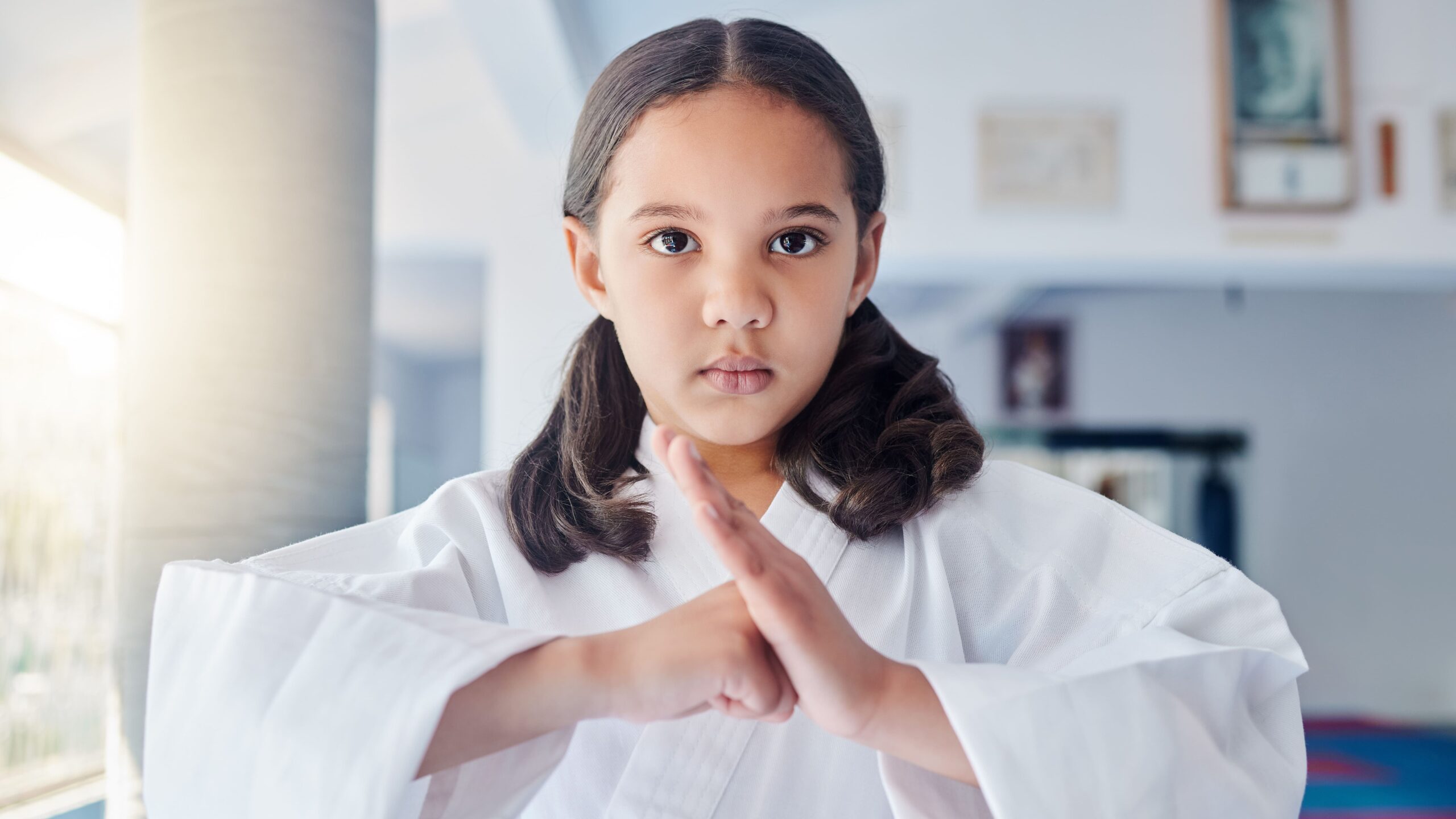 Menina usando quimono branco fazendo o gesto de saudação tradicional de respeito, encostando o punho na palma da mão dentro do dojo.