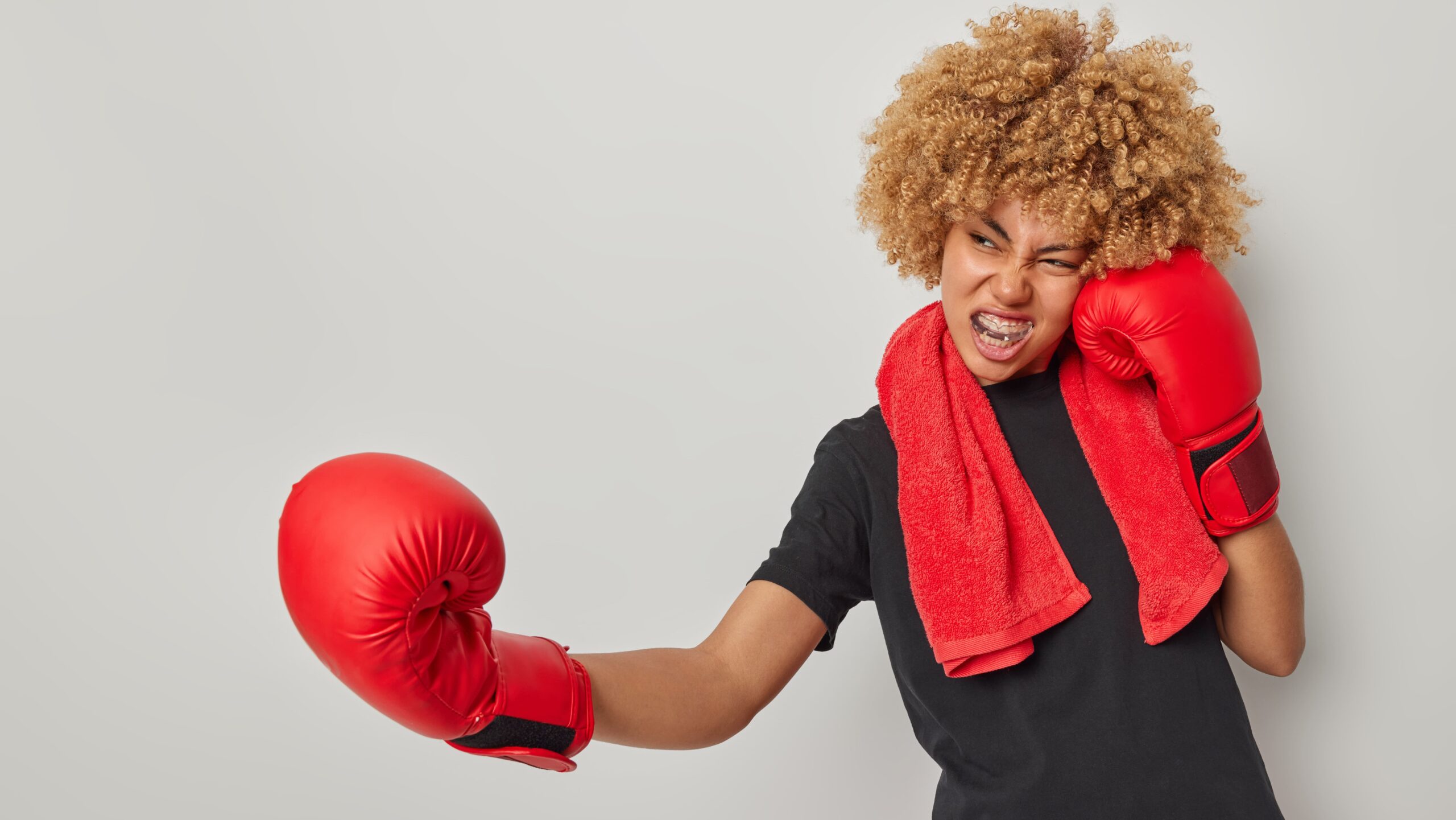 Mulher de cabelo crespo loiro com expressão de cansaço após treino, usando luvas de boxe vermelhas e toalha vermelha no pescoço.