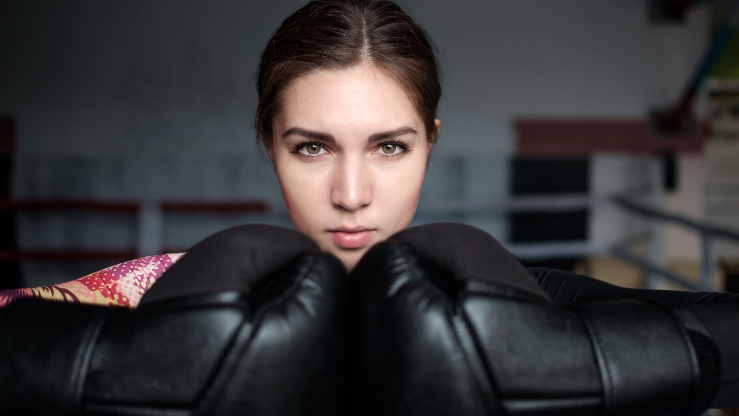 Mulher jovem dentro do ringue com olhar focado, segurando um par de luvas de boxe pretas na altura do rosto.