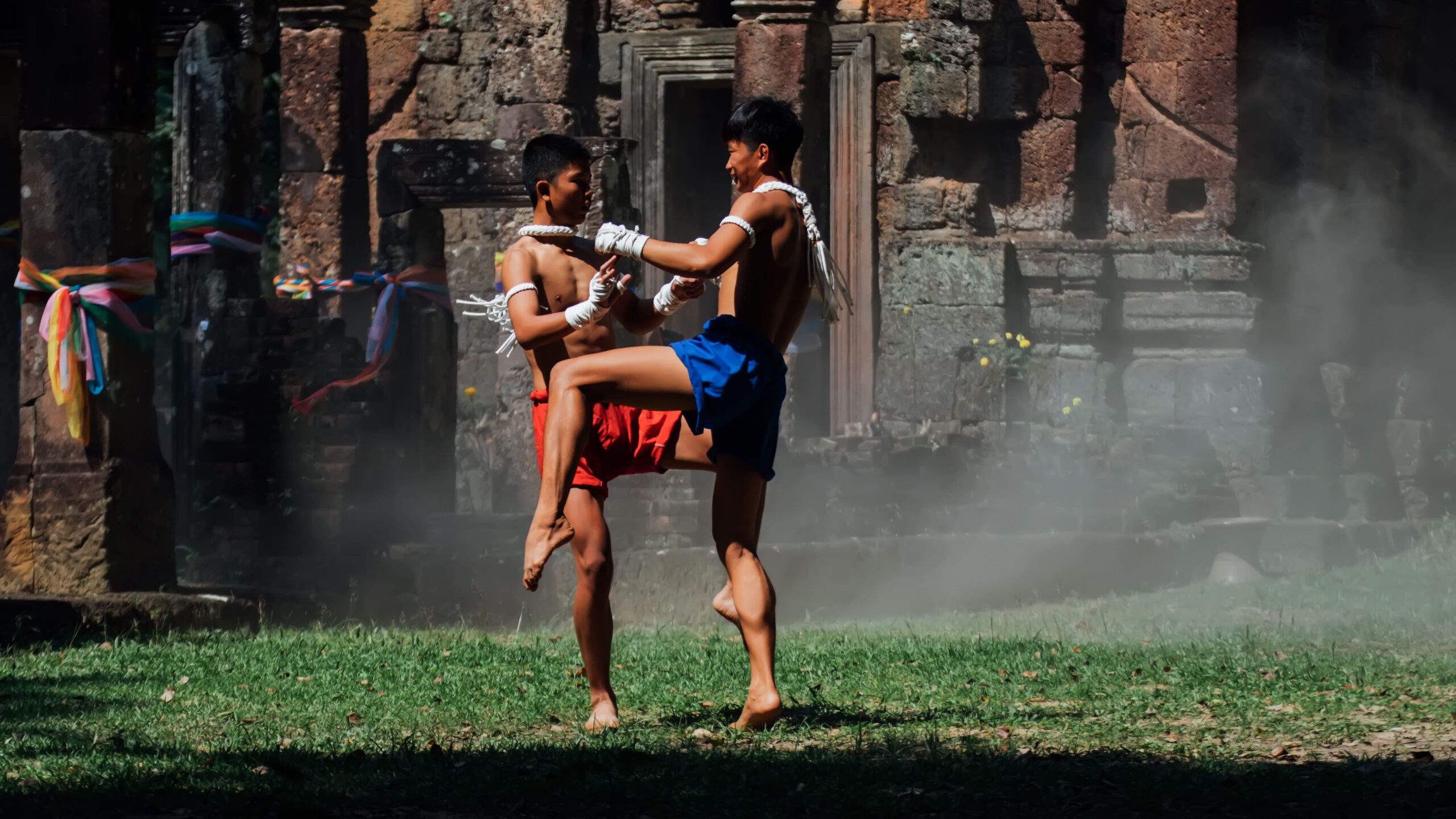 Dois praticantes de muay thai tradicional com mãos atadas treinando ao ar livre em frente a ruínas históricas. O lutador de calção azul avança elevando o joelho e estendendo os braços para buscar a pegada do clinch contra o oponente de calção vermelho.
