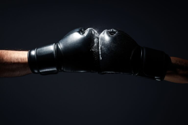 Duas pessoas em punhos fechados usando luvas de boxe pretas, uma fotografia em fundo escuro com luz dramática no centro, sugerindo combate e desafio.