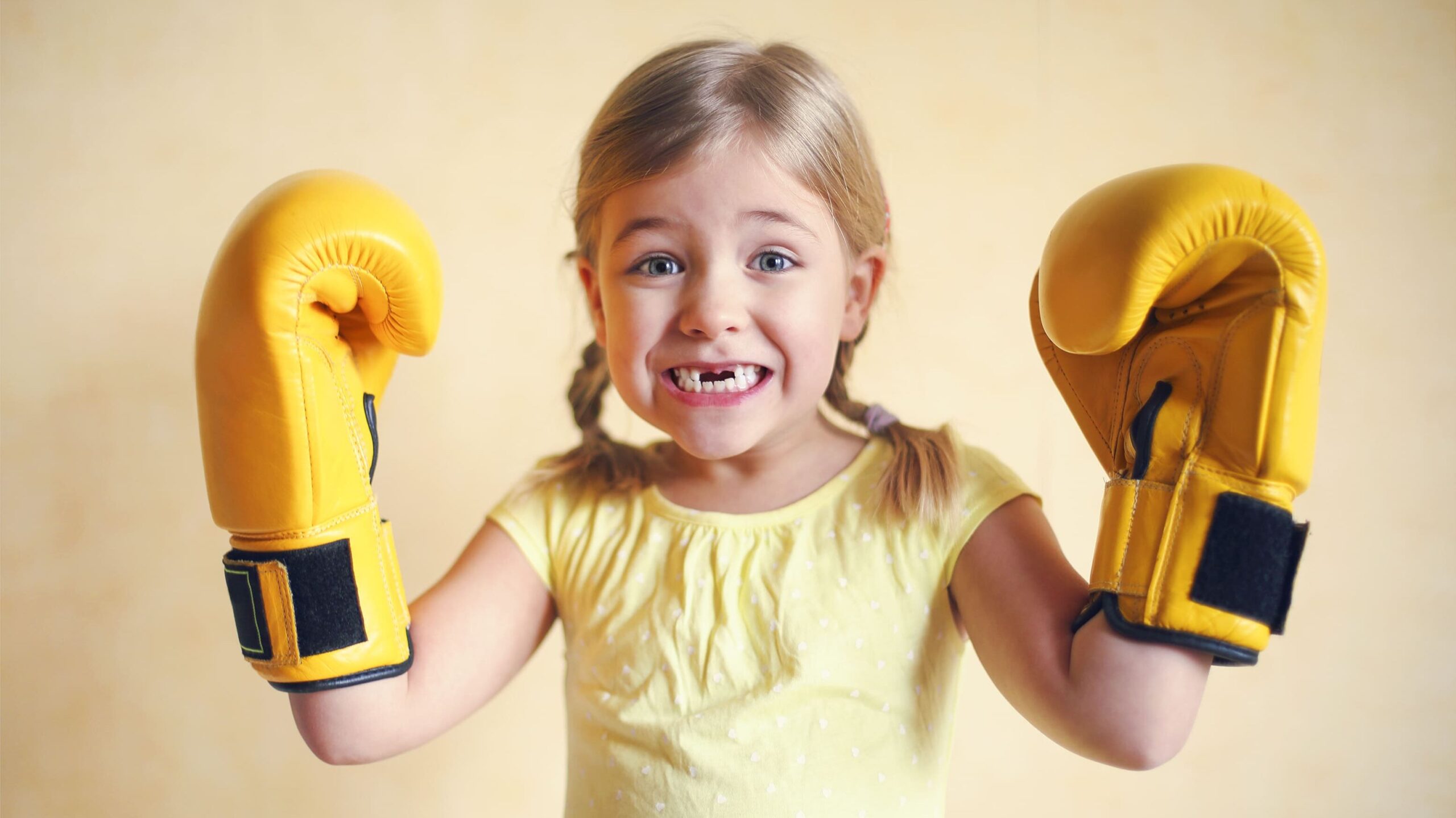 Menina sorridente vestindo camisetas amarela e usando luvas de boxe amarelas grandes, erguendo os braços de forma divertida.